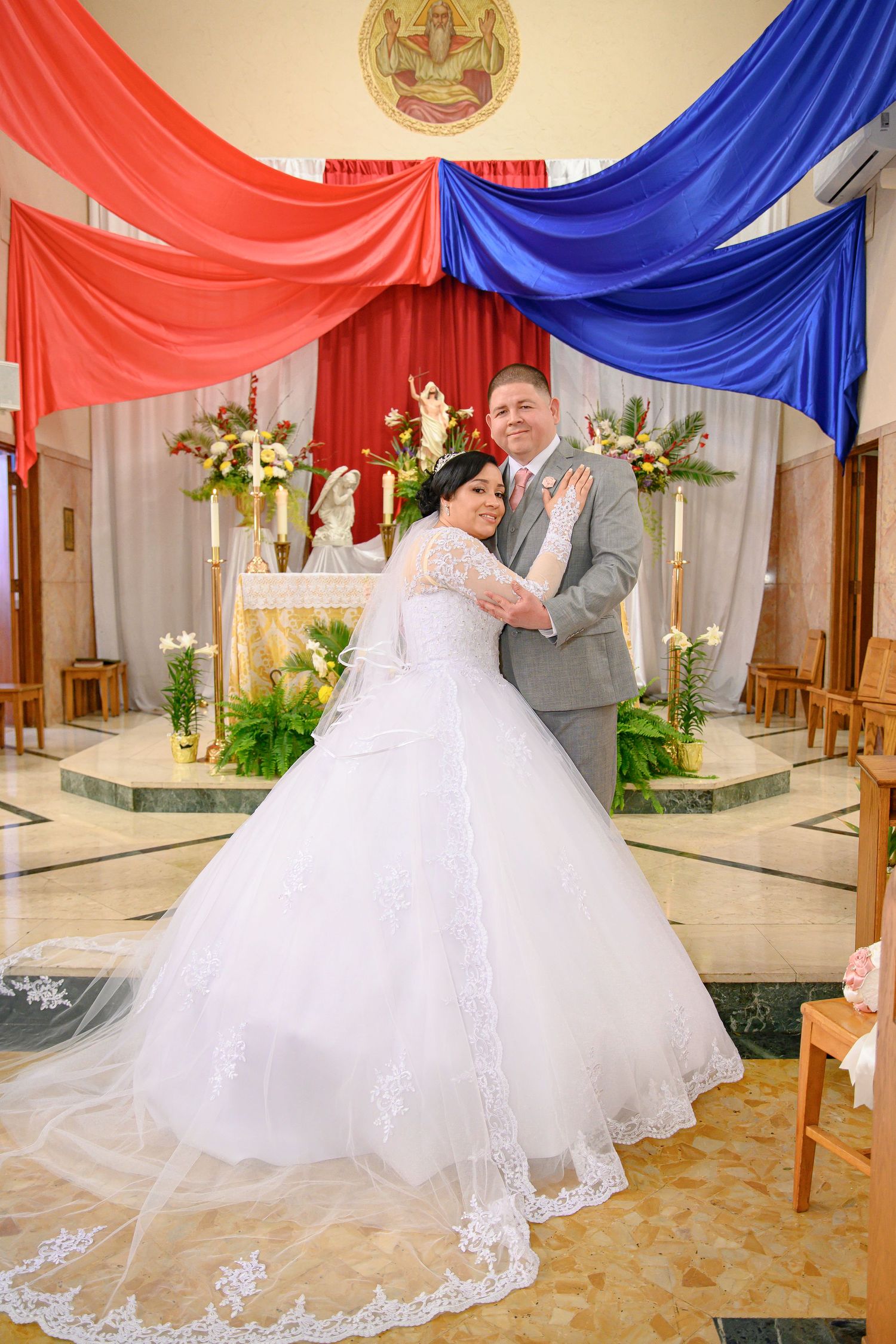 A couple shares an intimate moment during their wedding ceremony under red and blue draped fabric in a church setting.