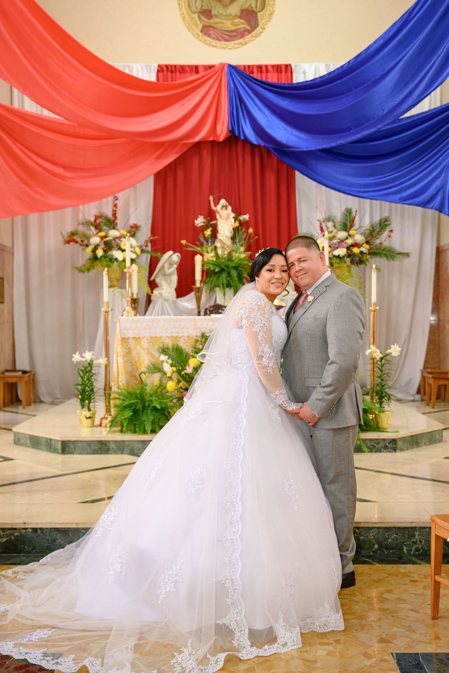 A wedding ceremony setup with red and blue draped fabric, white floral arrangements, and a decorated altar in a church setting.