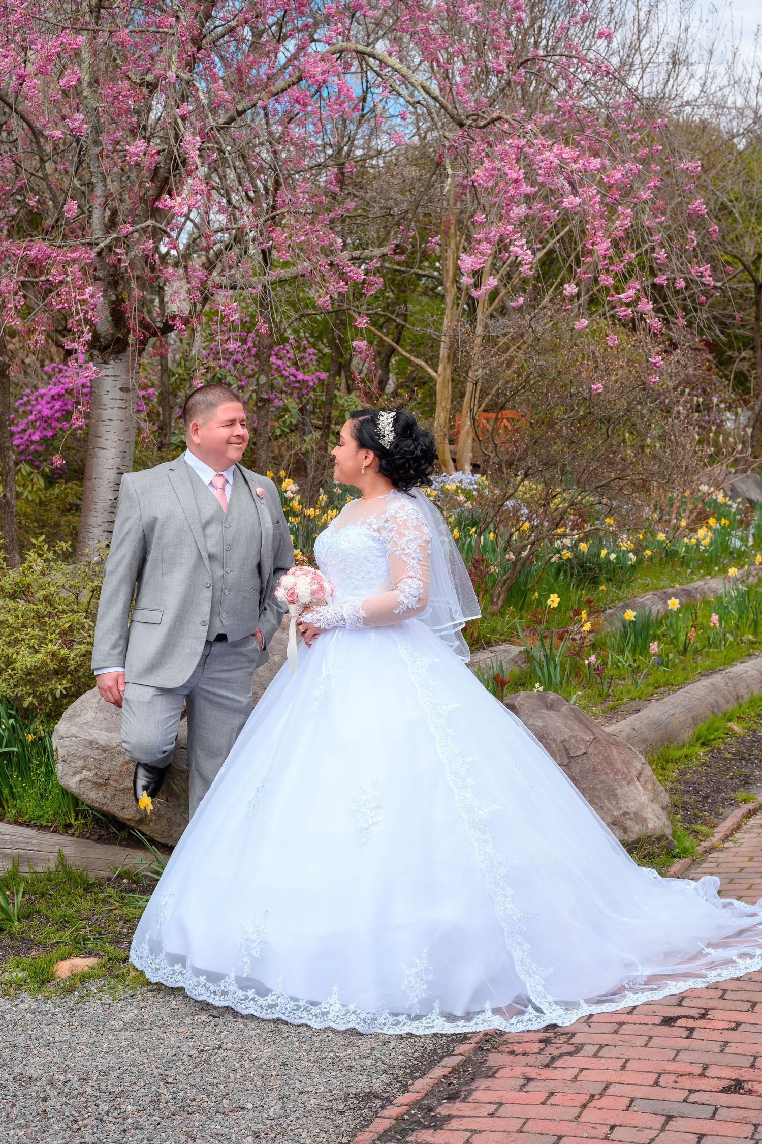 A couple in wedding attire poses beneath blooming cherry blossoms on a brick path surrounded by spring flowers.