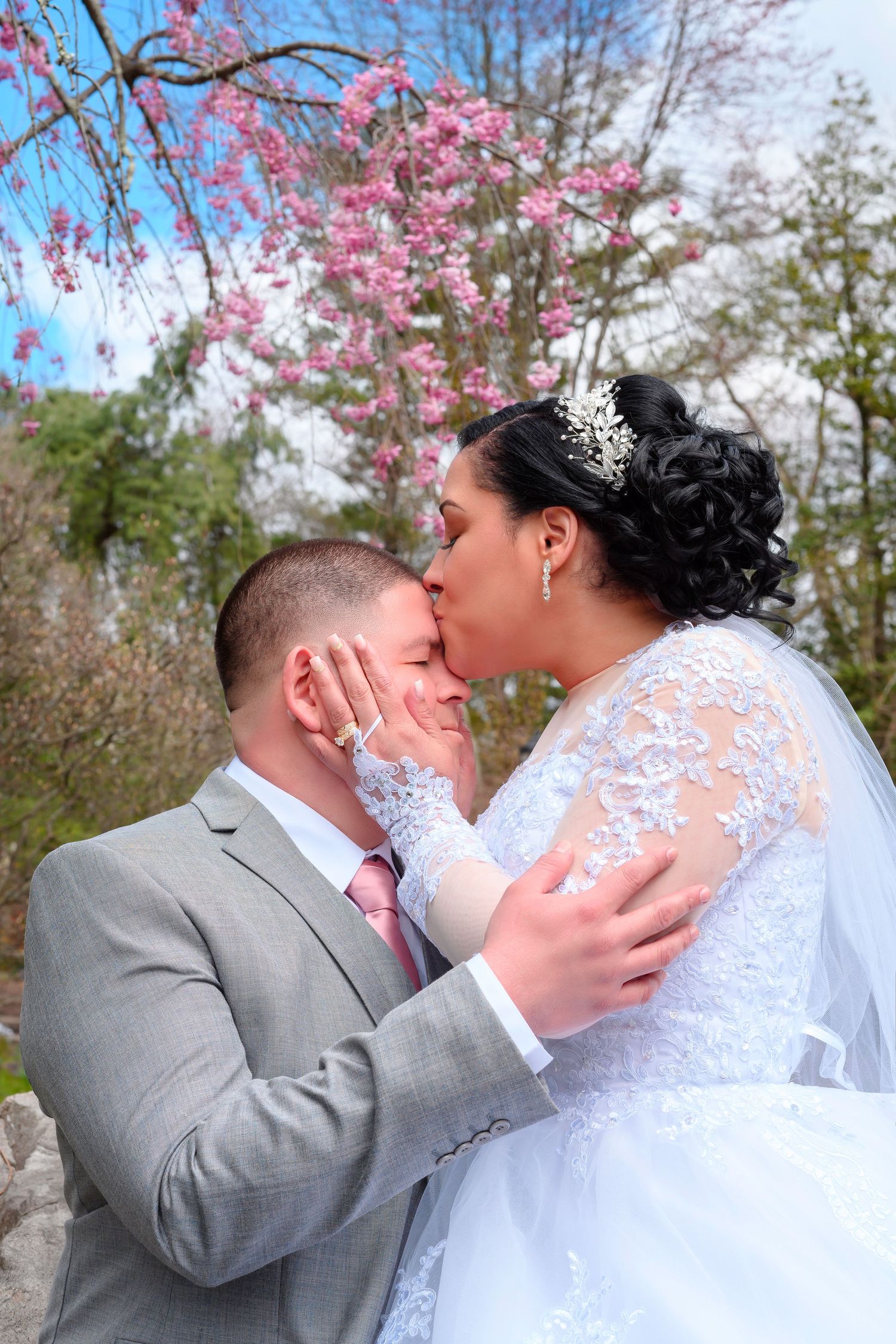A wedding couple shares a romantic kiss under blooming cherry blossoms with the bride wearing a white lace gown.