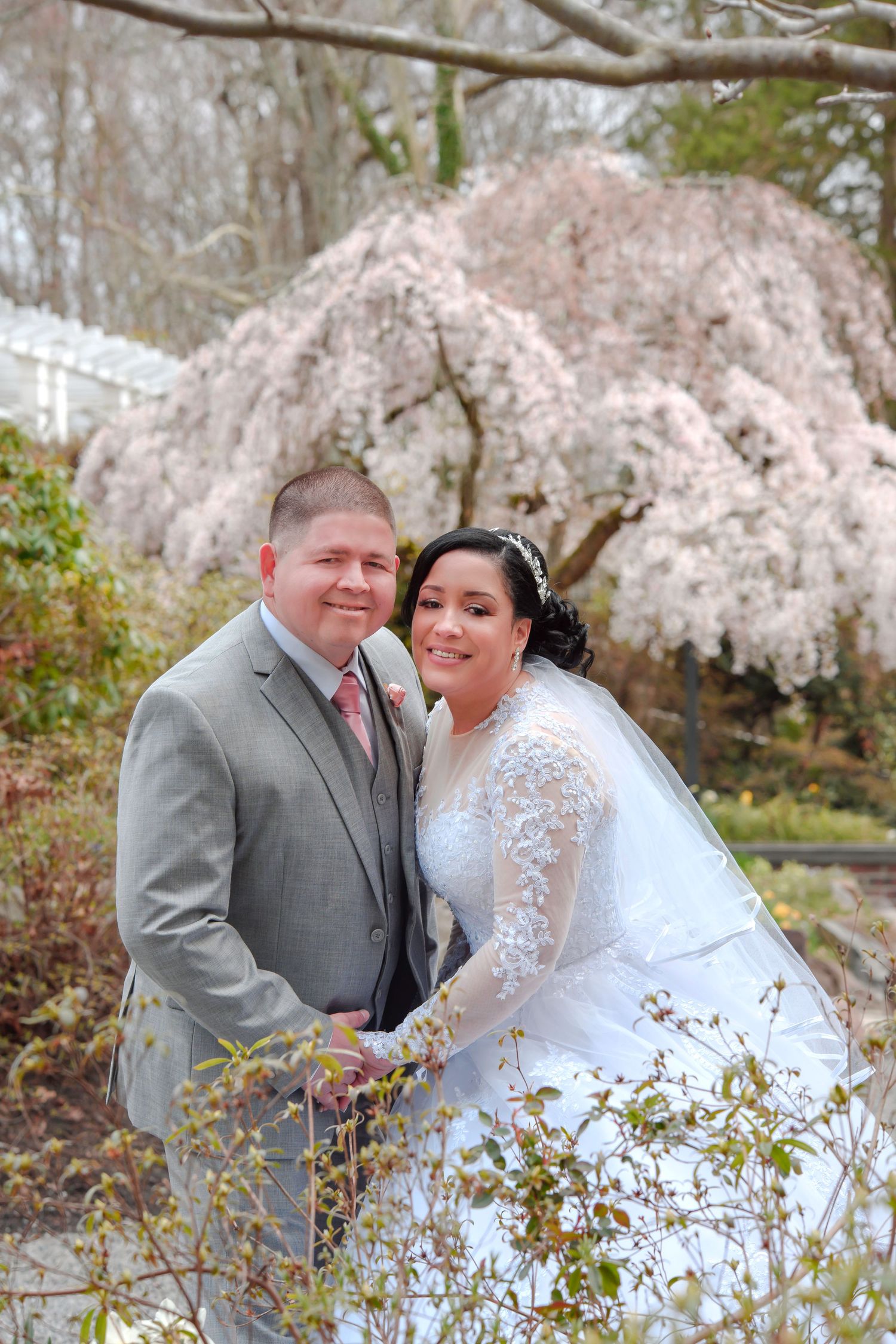 A couple poses together under blooming cherry blossoms in a spring garden.