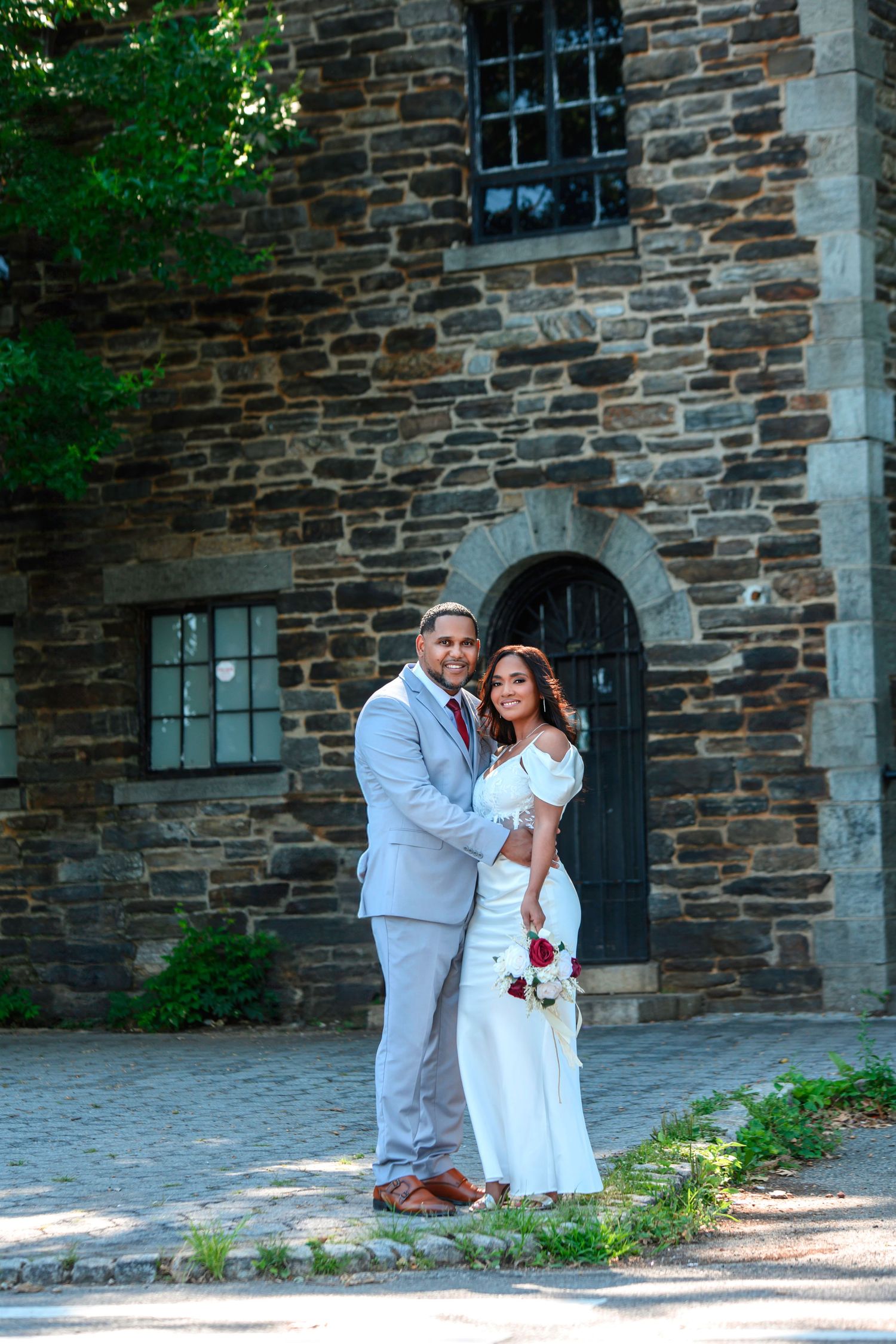 A couple poses together in wedding attire outside a historic stone church building with an arched doorway.
