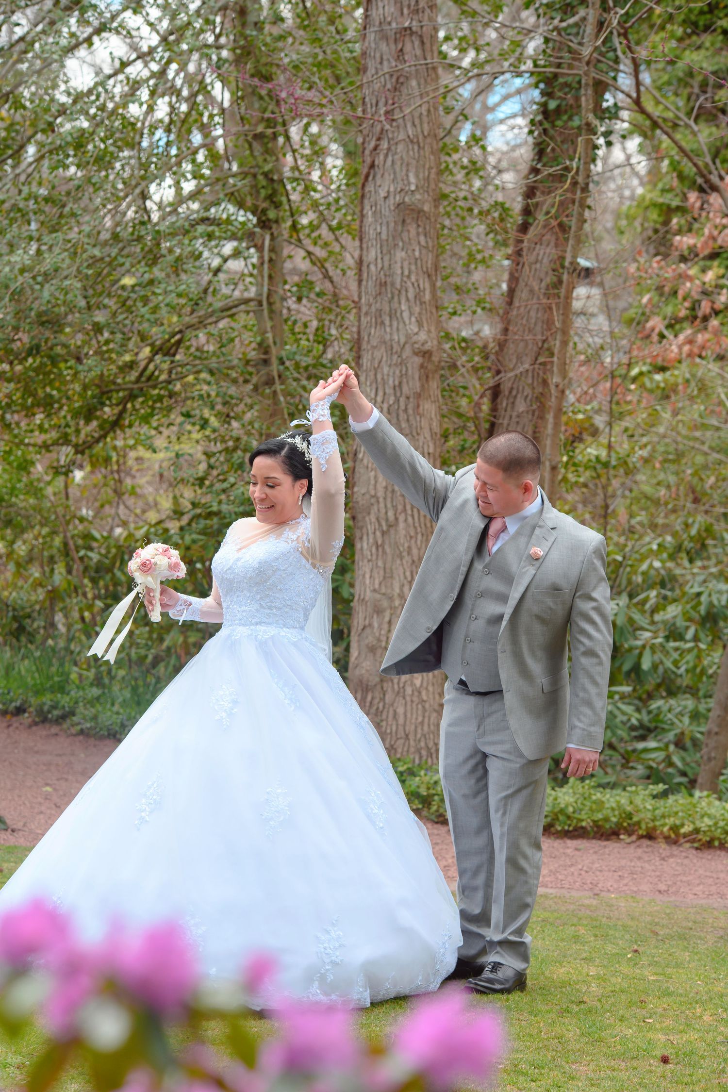 A couple celebrates their wedding day outdoors with a twirl on a garden path surrounded by trees and pink flowers.