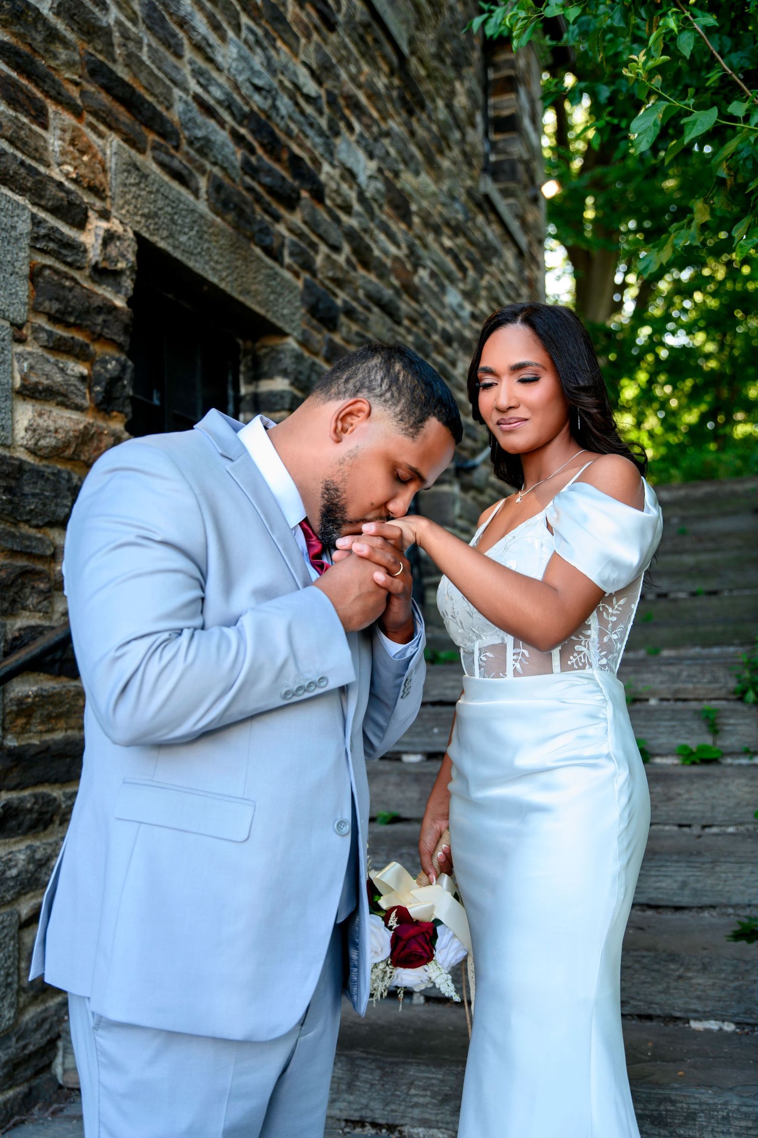 A romantic moment between newlyweds in light gray suit and white wedding dress against a stone wall backdrop.