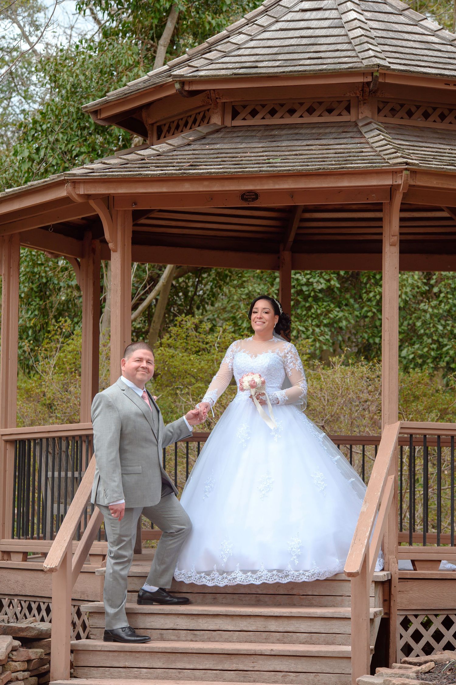 A couple poses in a rustic wooden gazebo with the bride in a white lace gown and groom in a gray suit.