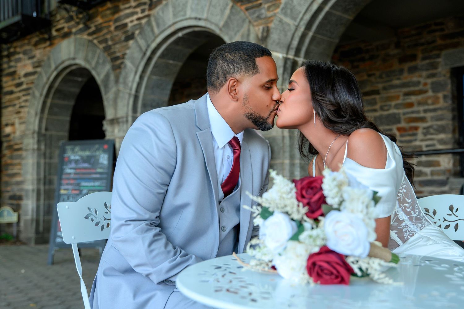 A romantic wedding moment at a stone archway with a bridal bouquet of red and white flowers on an elegant table.