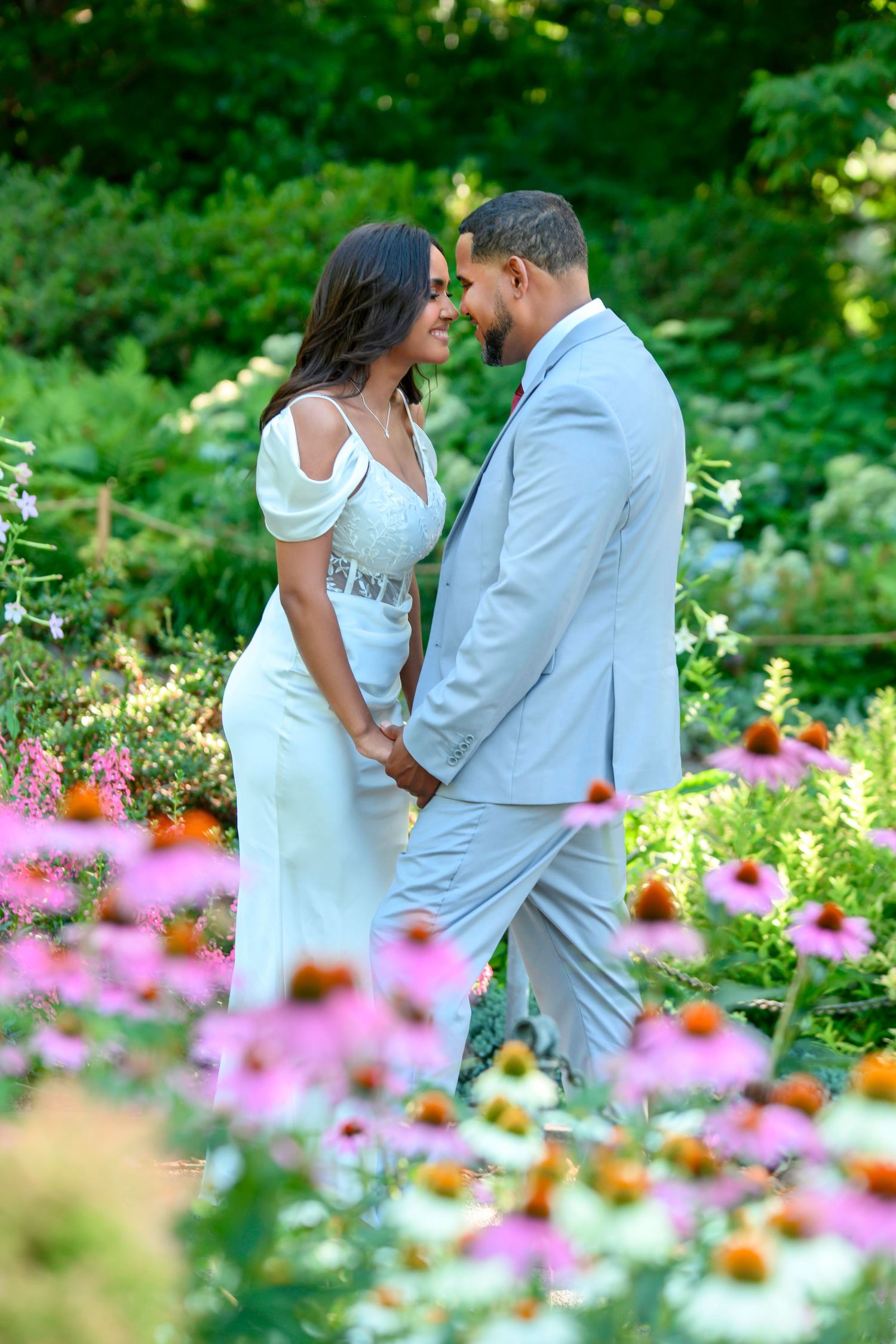 A romantic couple shares an intimate moment in a garden filled with vibrant pink and orange wildflowers on a sunny day.