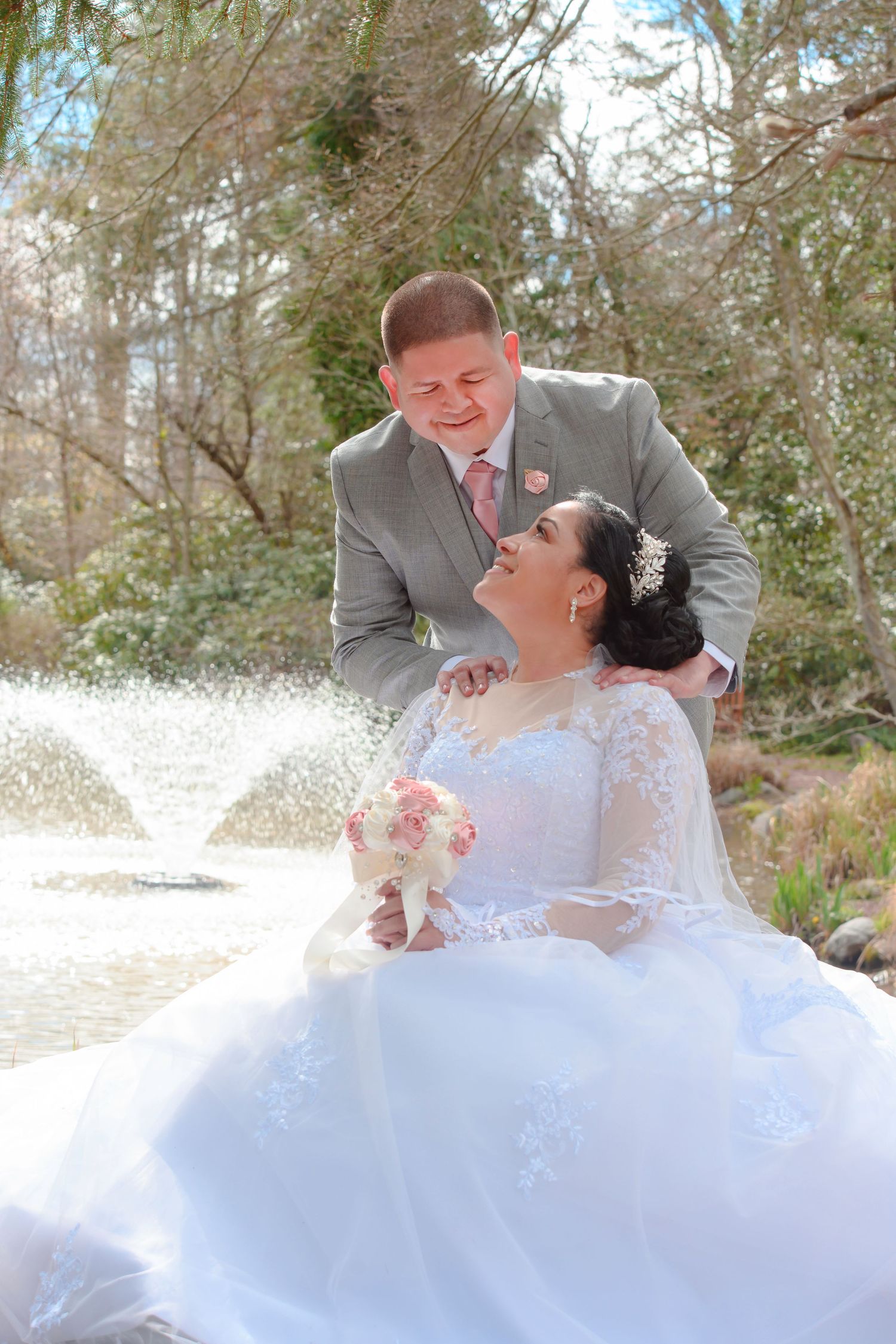 A happy couple shares a joyful moment outdoors on their wedding day, with the bride in a white gown holding a pink bouquet.