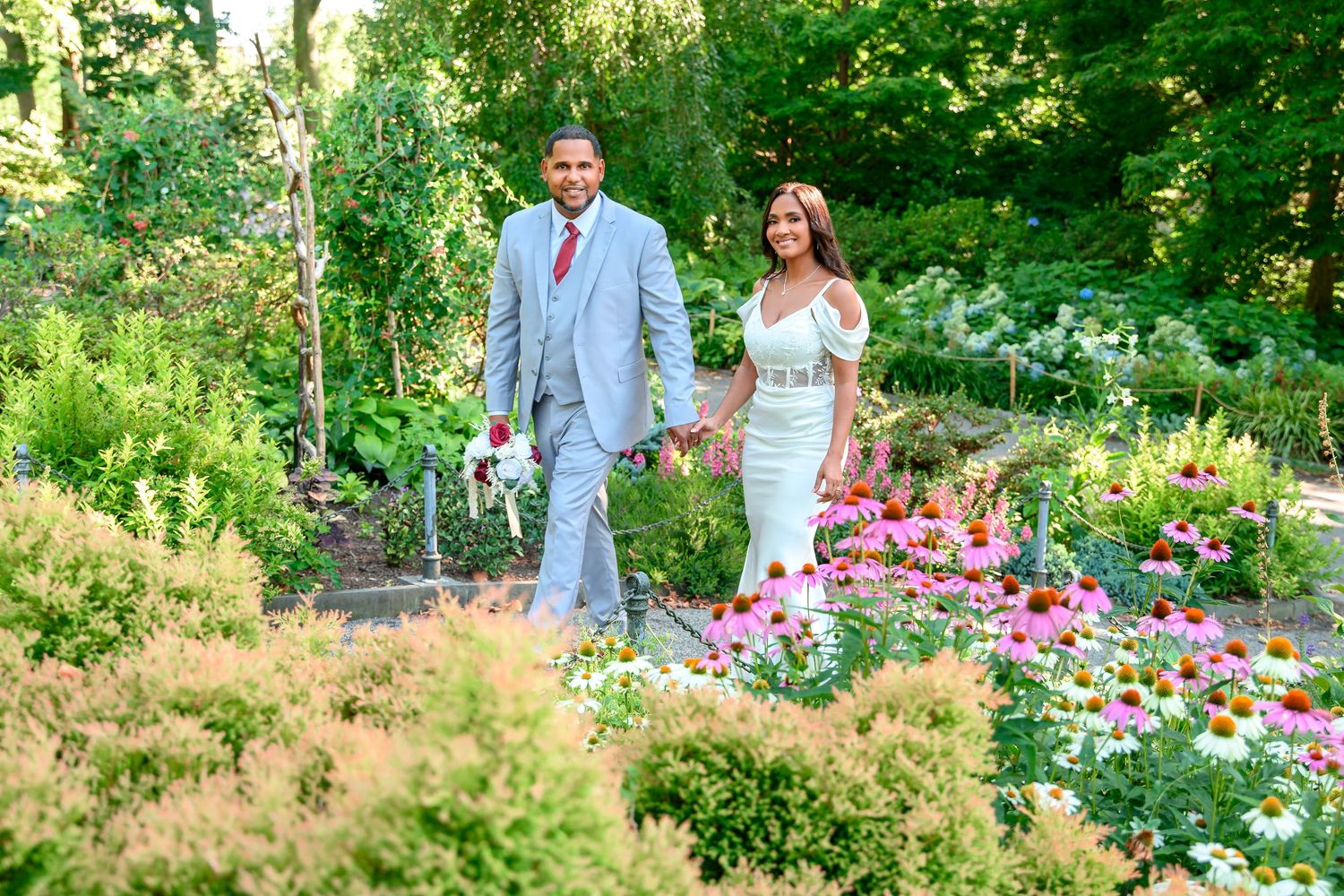 A couple walks together in a lush garden filled with vibrant pink flowers and greenery on a sunny summer day.