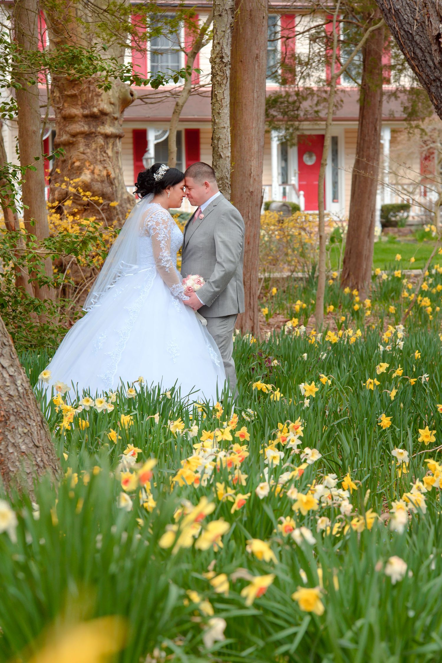 A couple stands together in a spring garden filled with blooming yellow daffodils in front of a pink house.