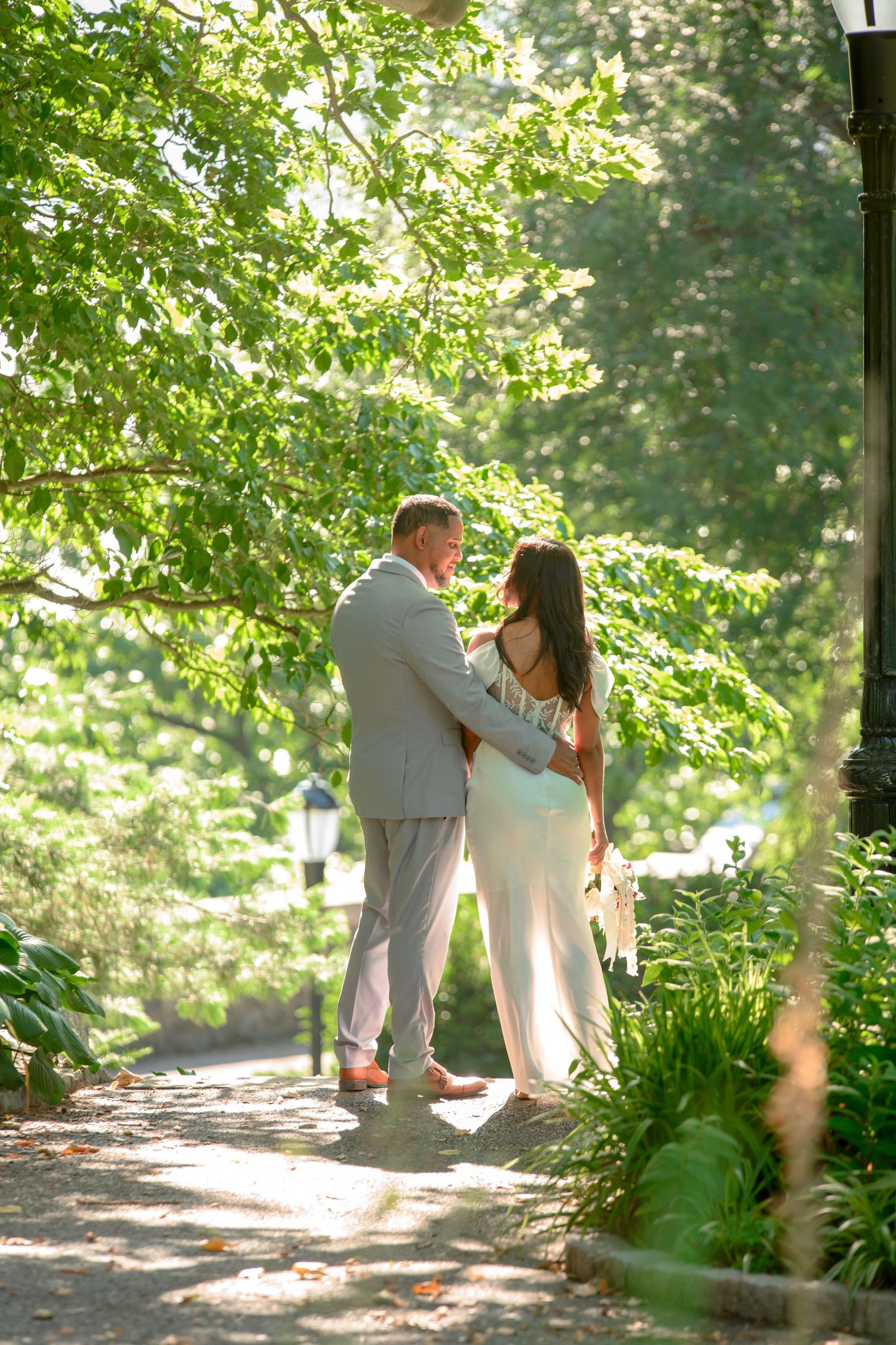A couple embraces on a sunlit garden path surrounded by lush green foliage and dappled sunlight.