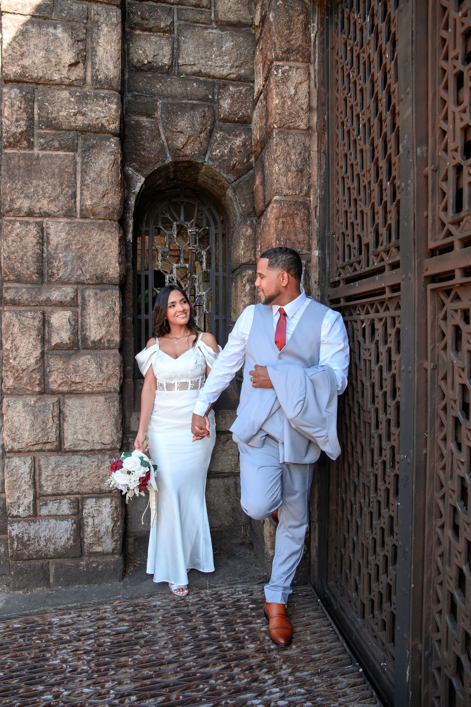 A wedding couple walks through a stone archway with ornate metal gates at a historic building.