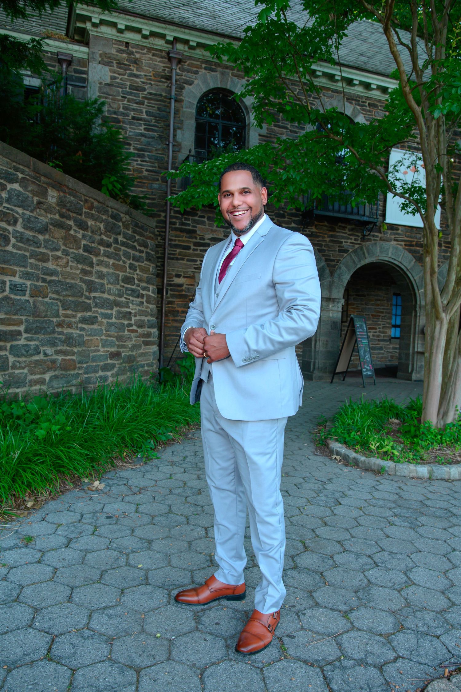 Someone poses in a light gray suit with brown shoes against a stone building with arched windows and green trees.