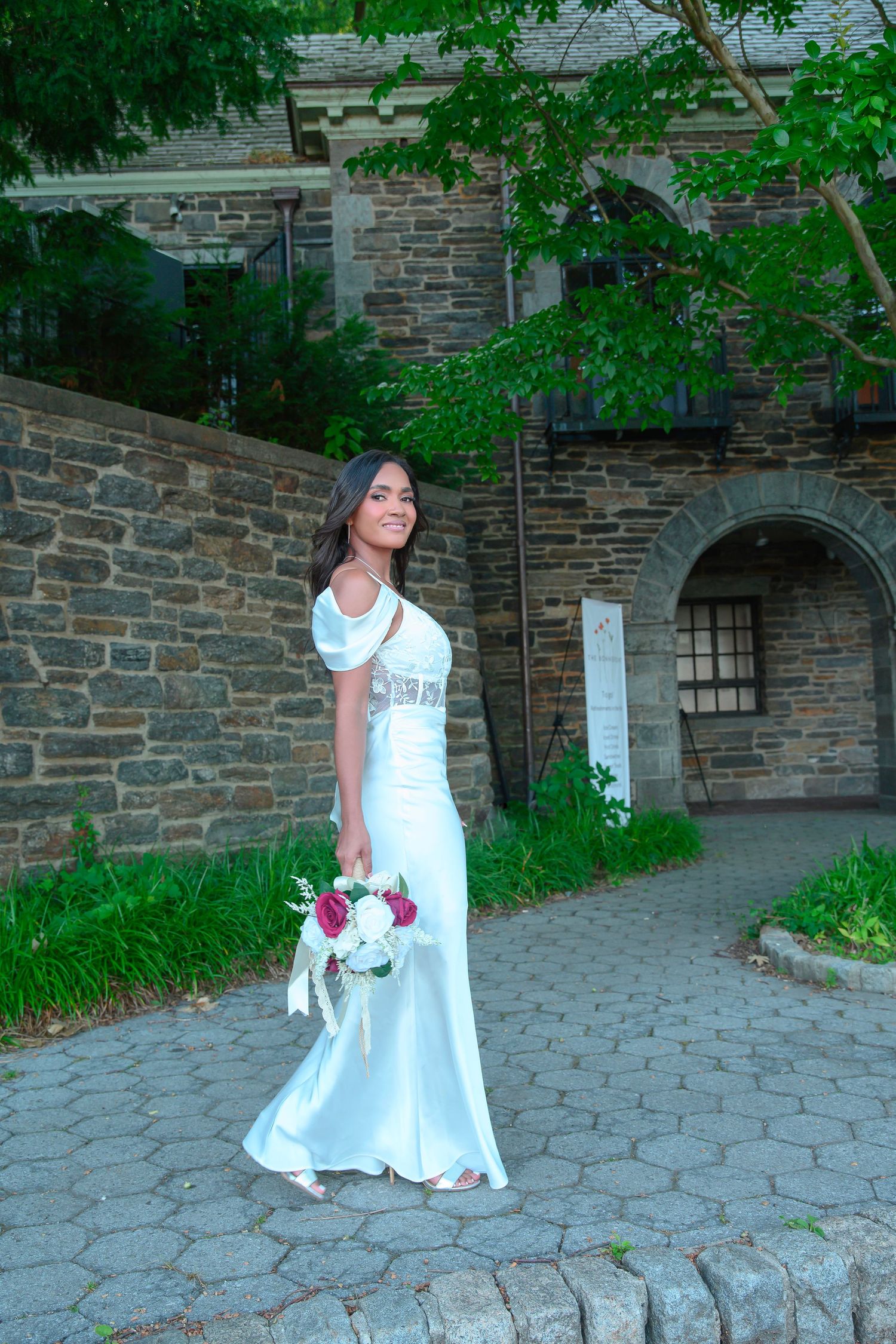 A person in an elegant white wedding gown holds a bouquet while posing against a stone wall archway surrounded by greenery.