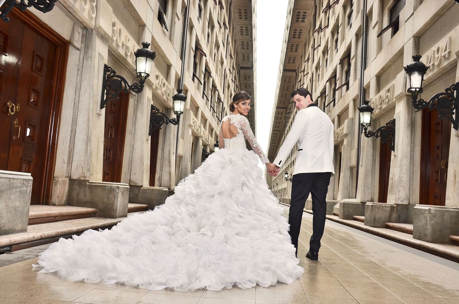 A couple in wedding attire walks hand in hand down a narrow historic street lined with elegant lampposts.