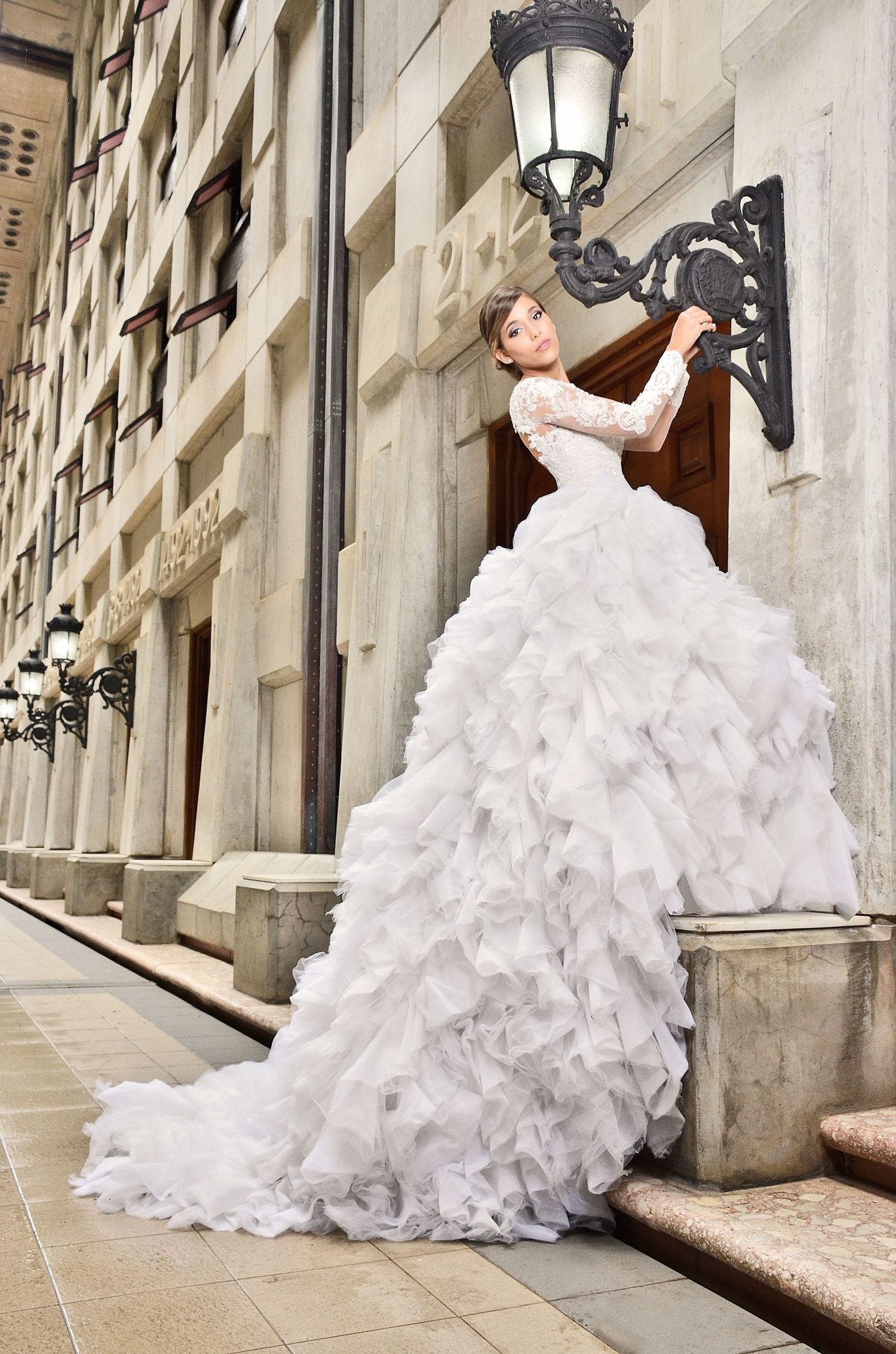 A bride in a dramatic ruffled white wedding gown poses against a classic stone building with vintage lampposts.