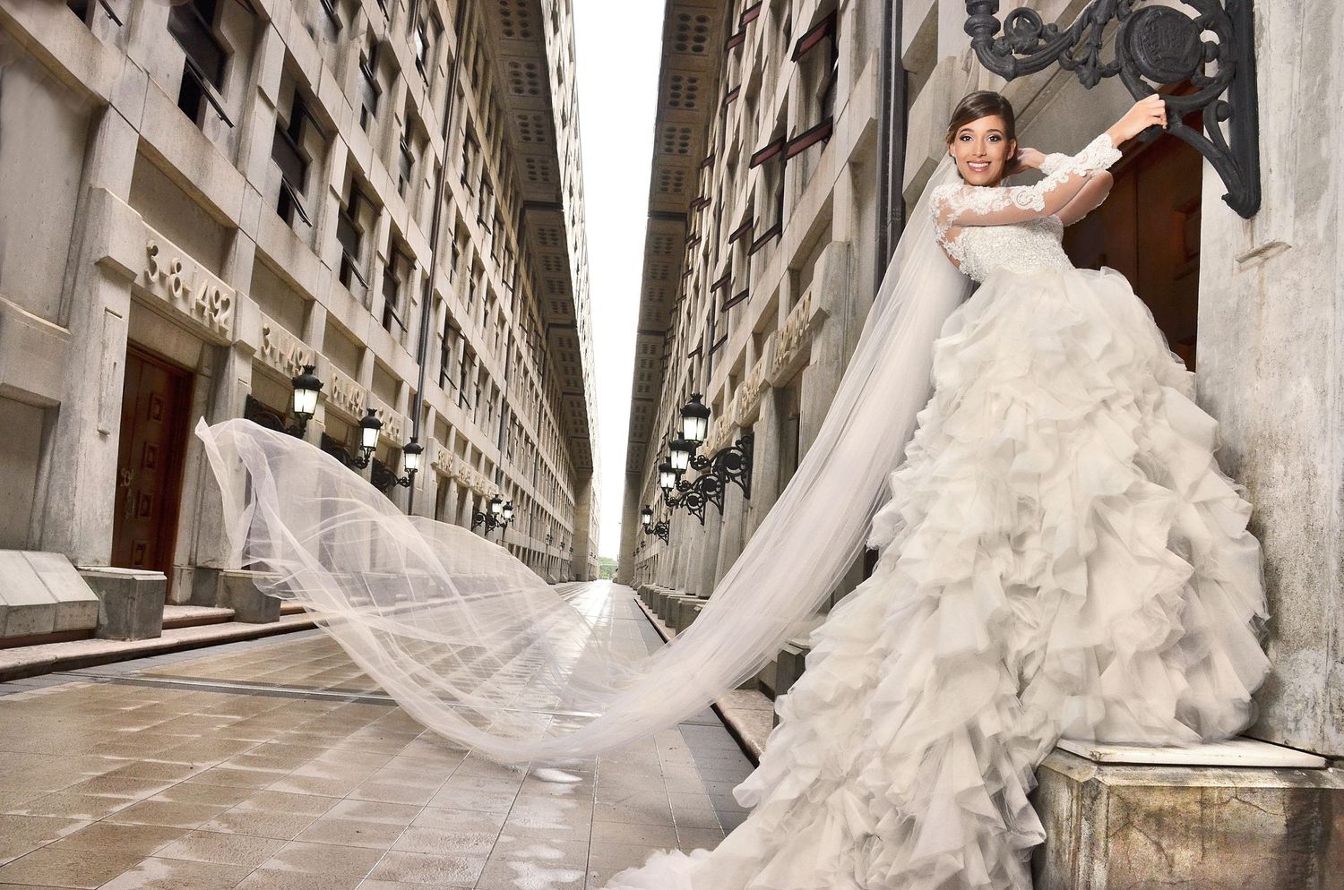 A flowing white wedding gown with dramatic ruffles and long veil creates a striking silhouette against stone architecture.