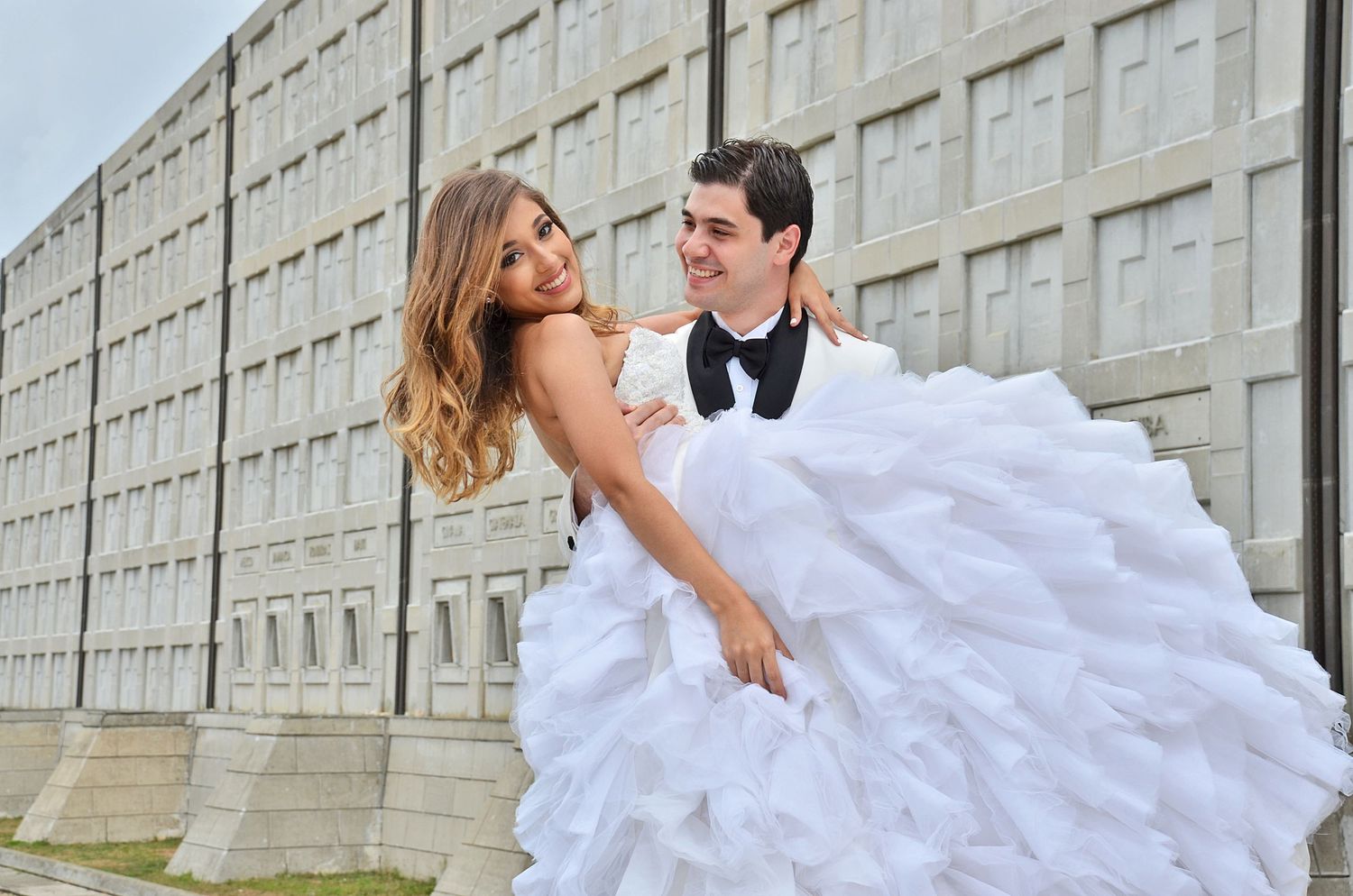 A bride in a voluminous ruffled white gown dips back while dancing with a groom in a black tuxedo in front of a concrete wall.