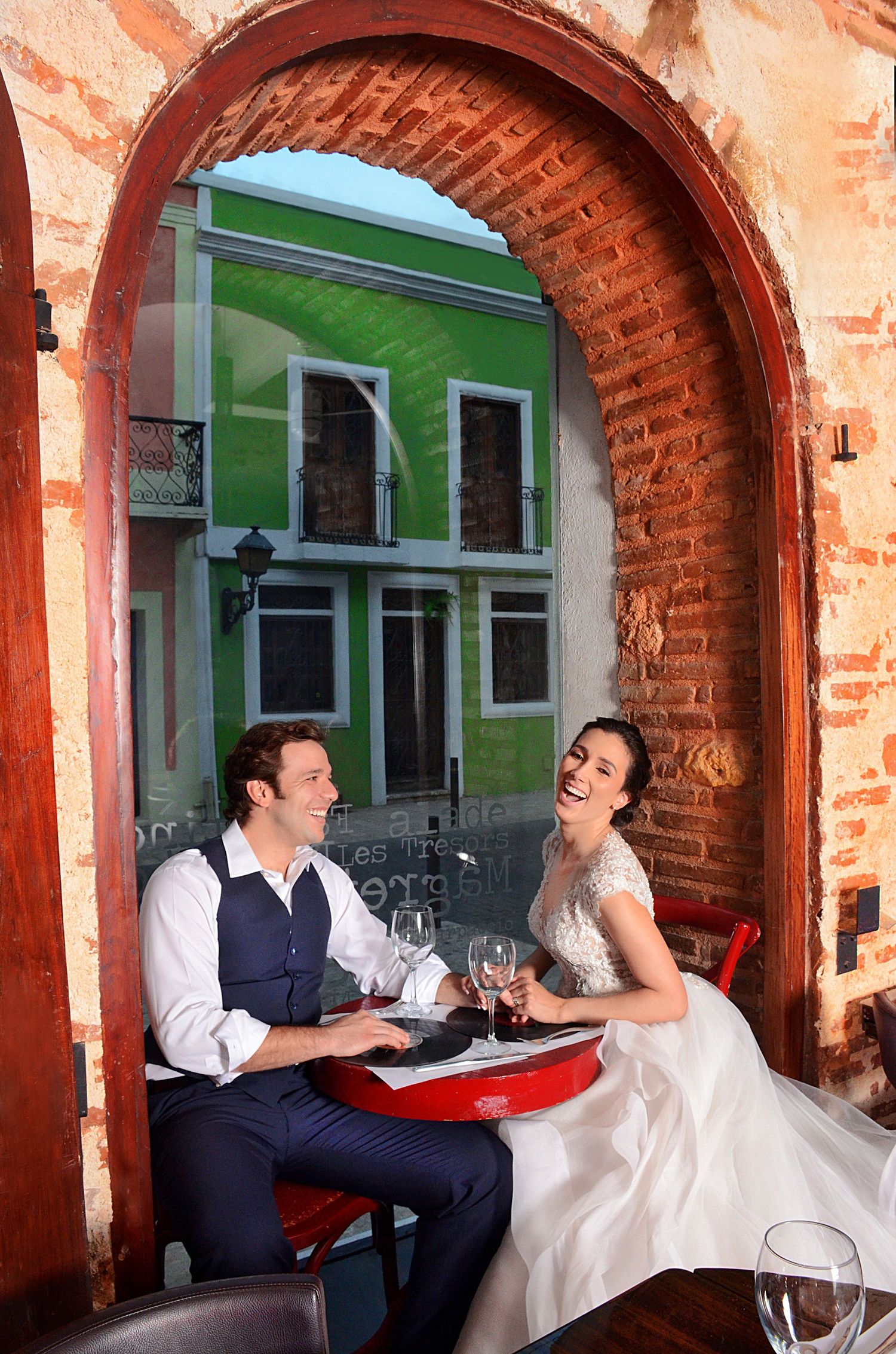 A couple shares wine and laughter at a table under a rustic brick archway with a bright green building in the background.