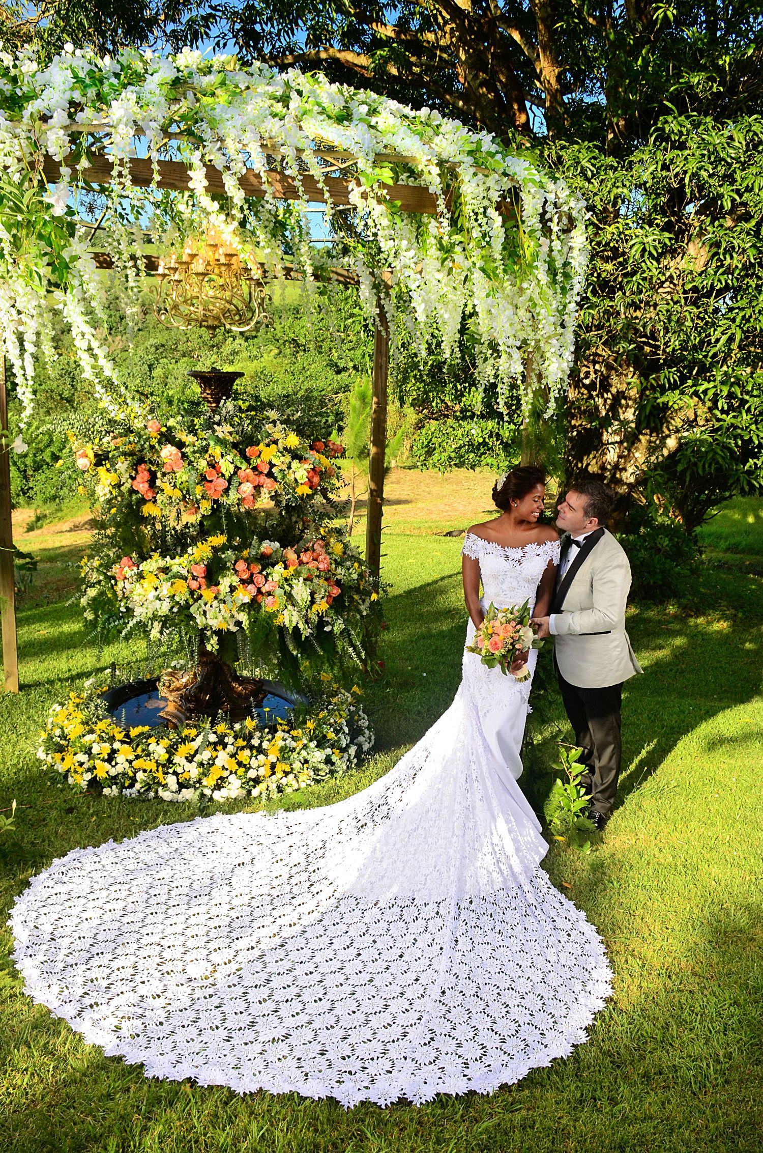 Romantic outdoor wedding scene with cascading wisteria, floral arch, and bride in flowing lace gown with dramatic train.