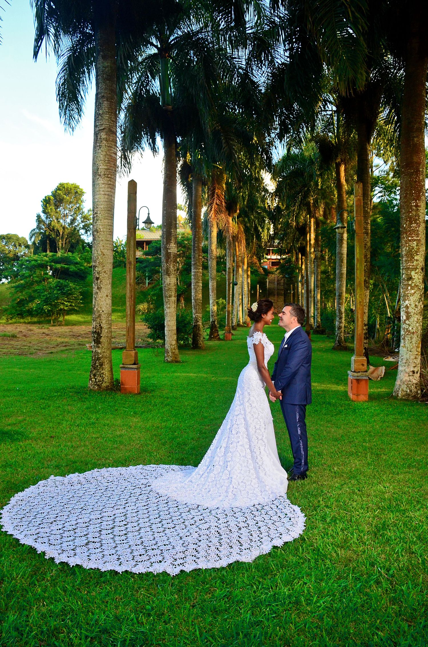 A wedding couple standing in a majestic palm tree-lined avenue with an intricate lace train on display.