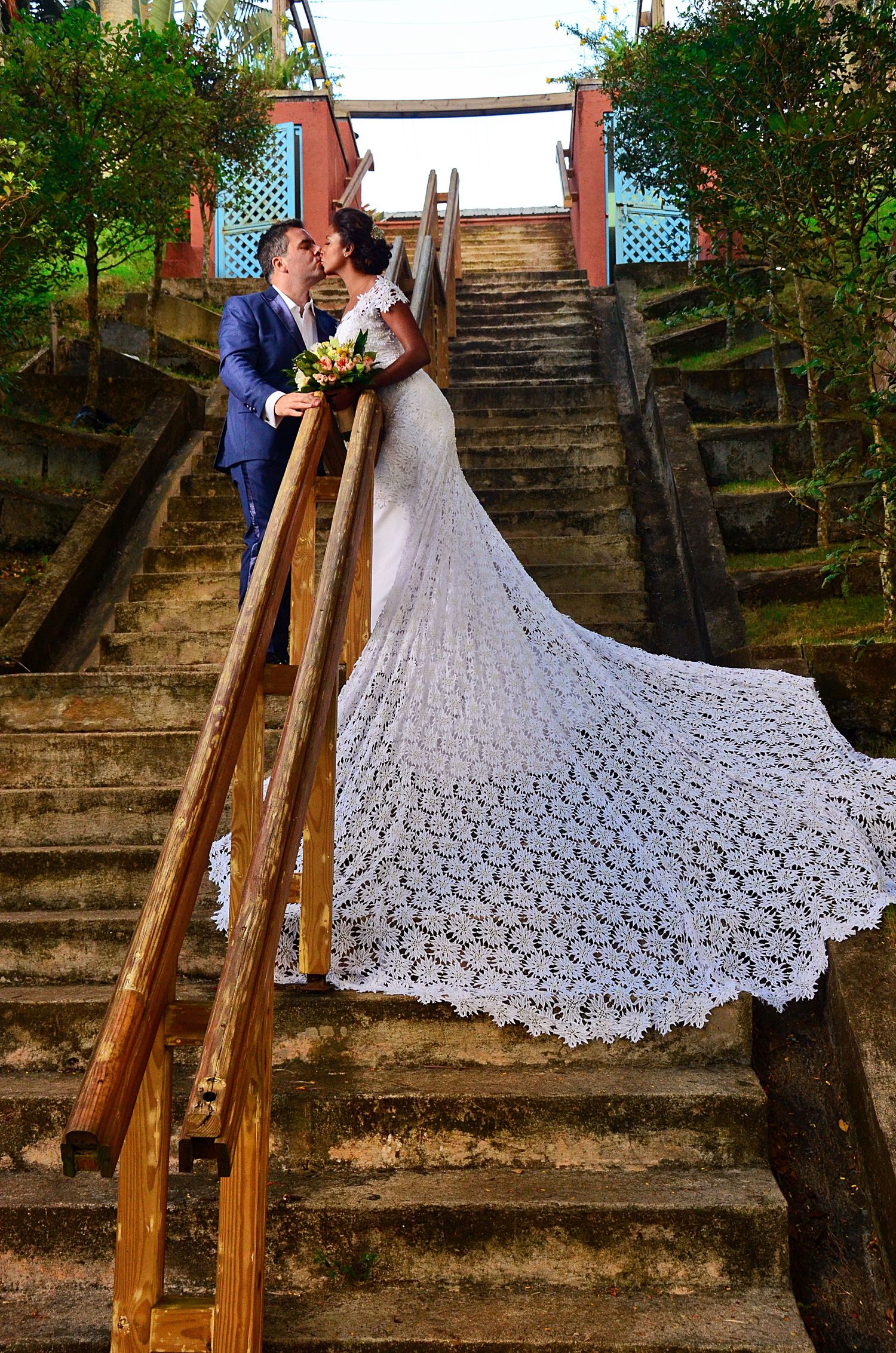 Wedding couple stands on rustic stone stairs with dramatic lace train flowing down the steps.