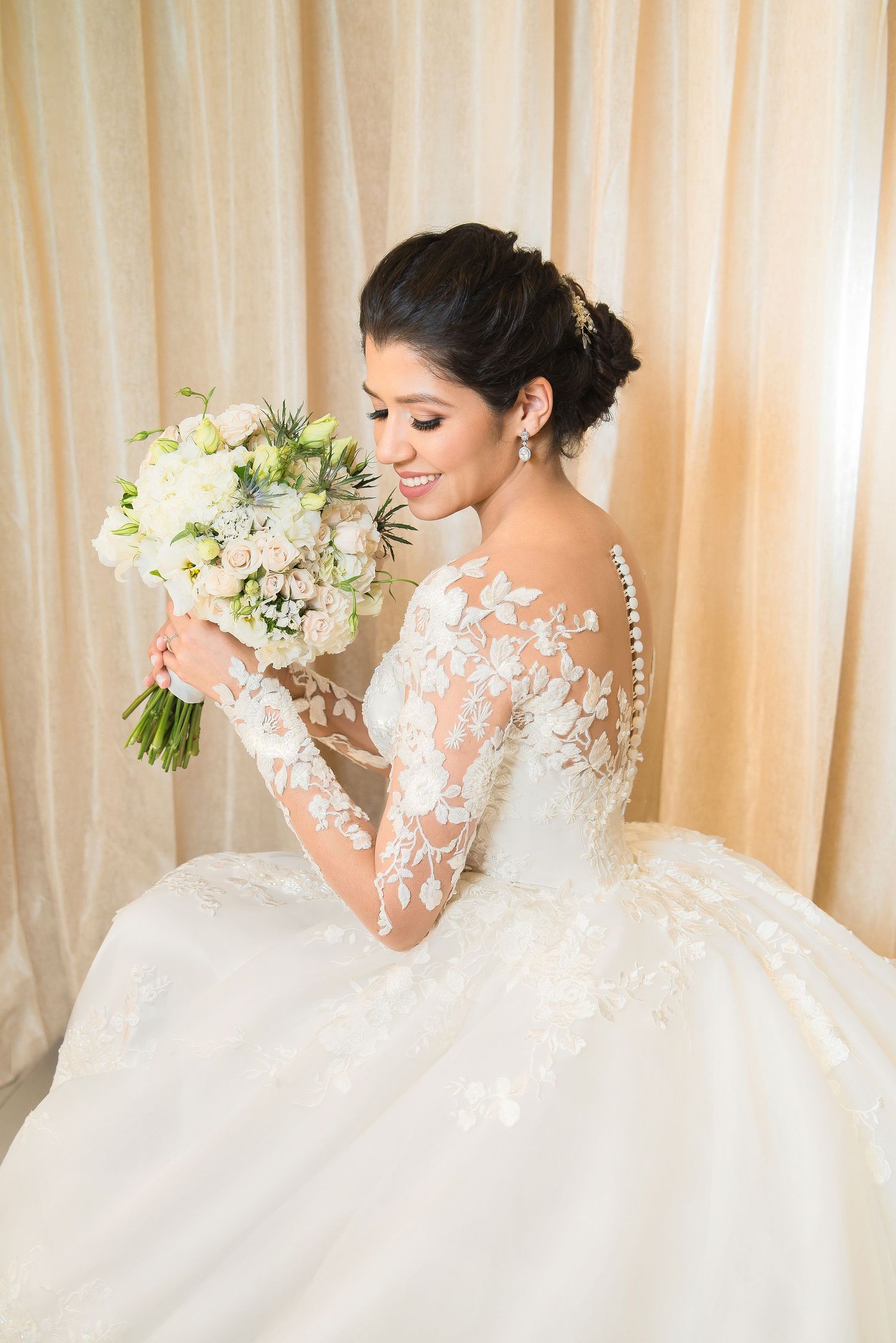 Bride in elegant lace long sleeve wedding gown holds white floral bouquet while smiling against cream backdrop.