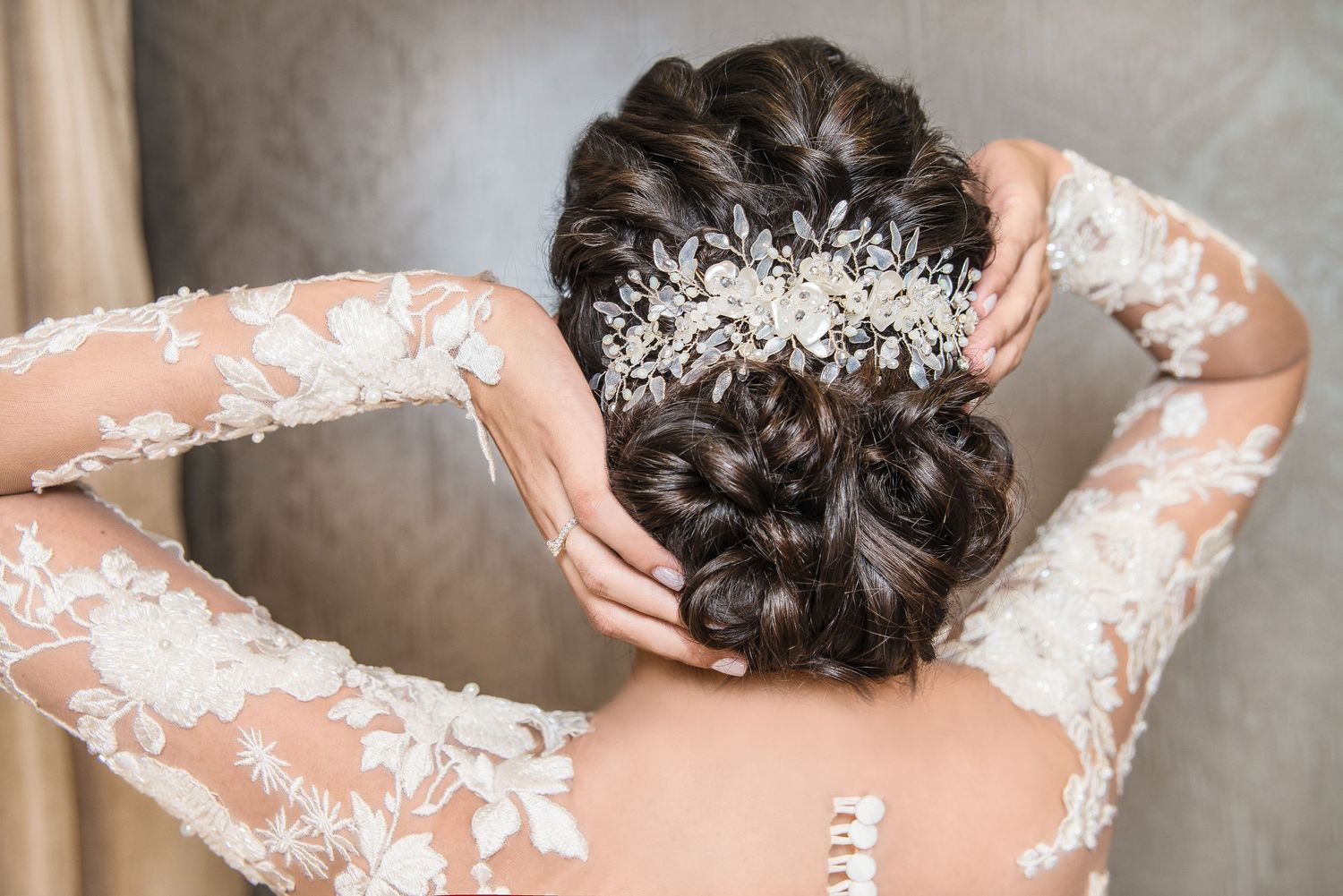 Elegant bridal updo hairstyle with ornate crystal hair accessory and delicate lace sleeve detail.