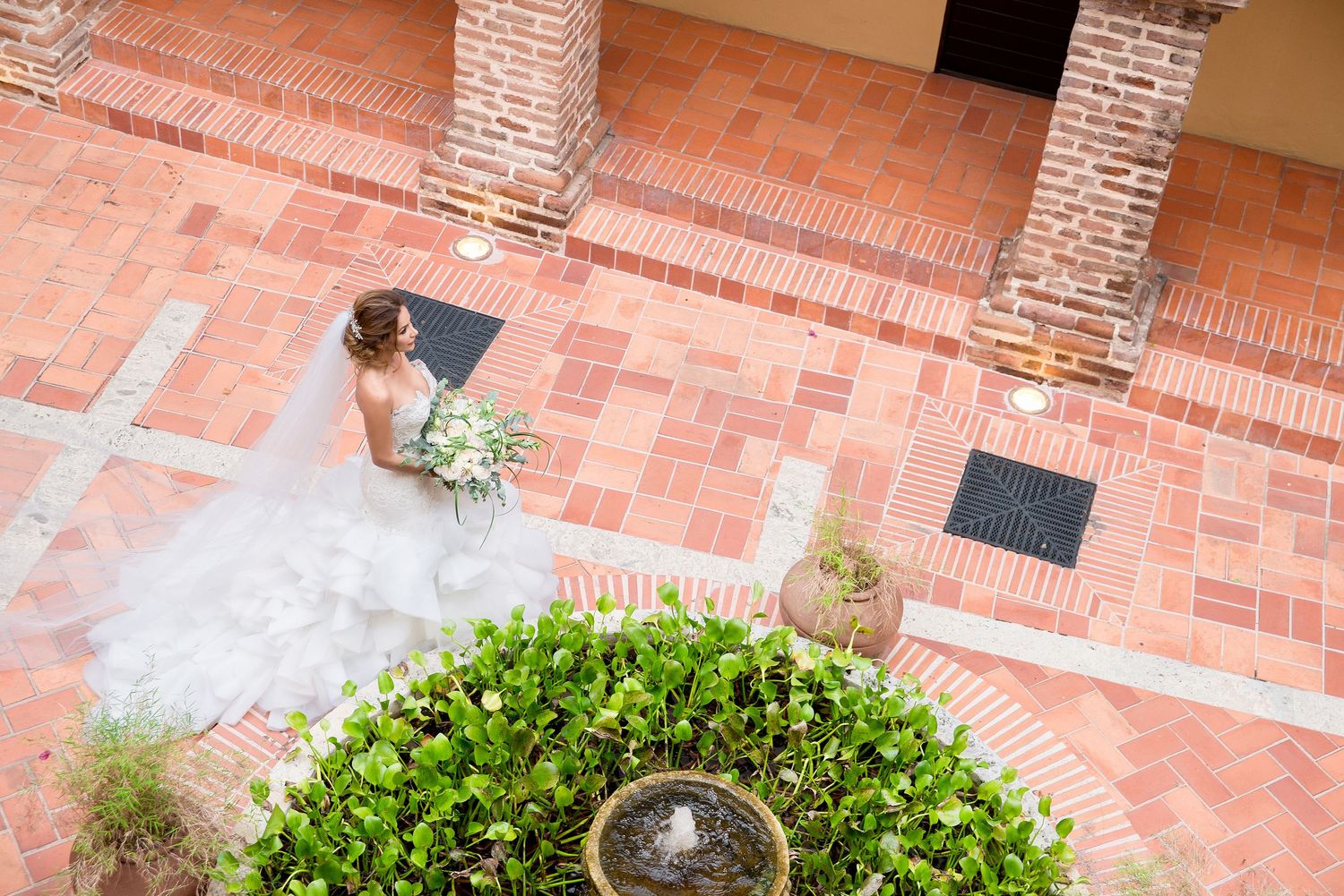 Overhead view of bride in white gown standing near fountain on terracotta tiled courtyard.