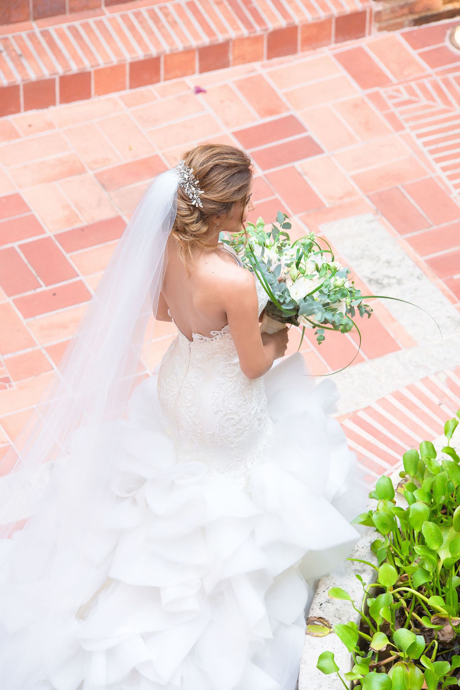 Bride in flowing white wedding gown holds a green and white bouquet while standing on red brick steps.