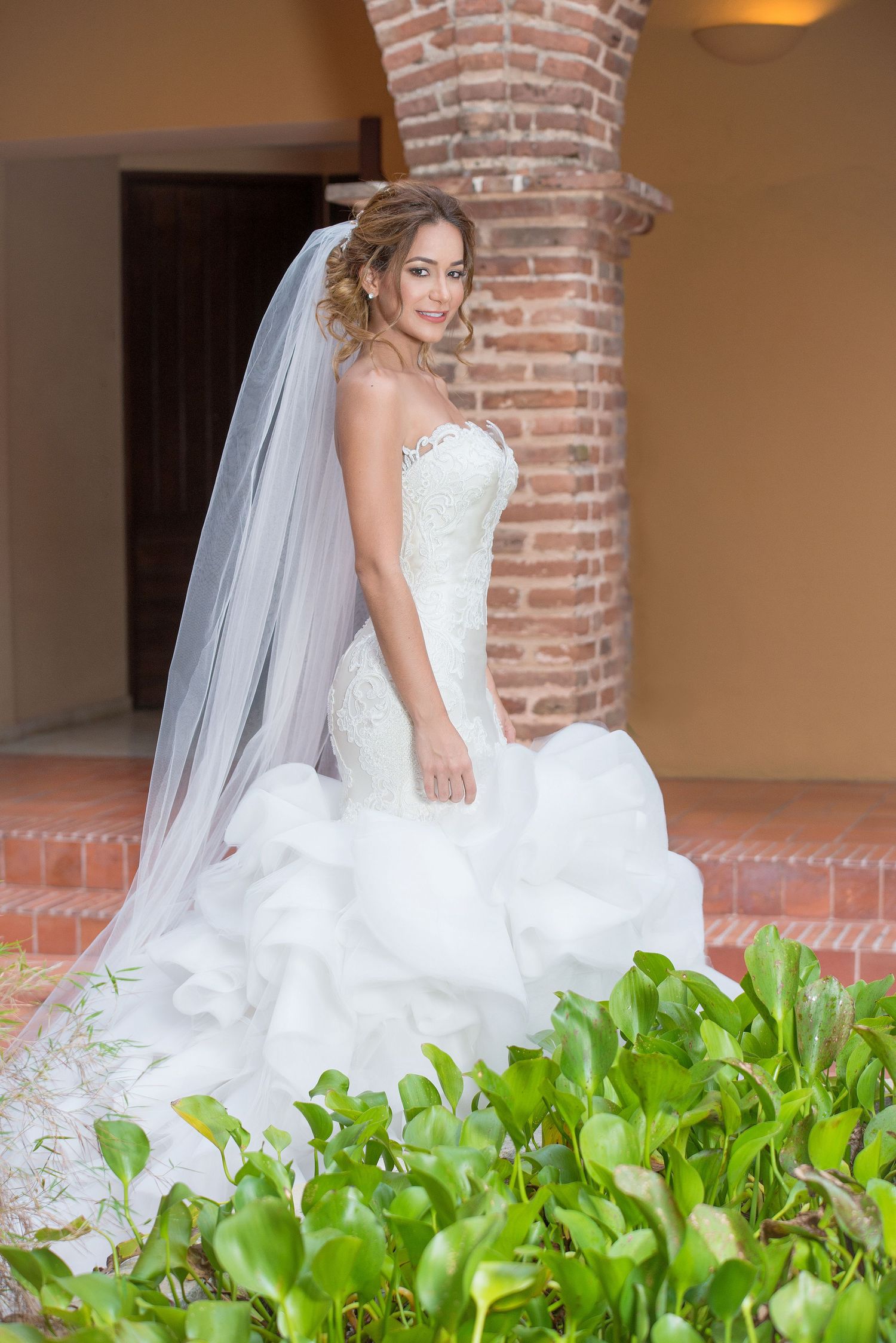 A wedding dress with a ruffled mermaid silhouette displayed against exposed brick architecture and green foliage.