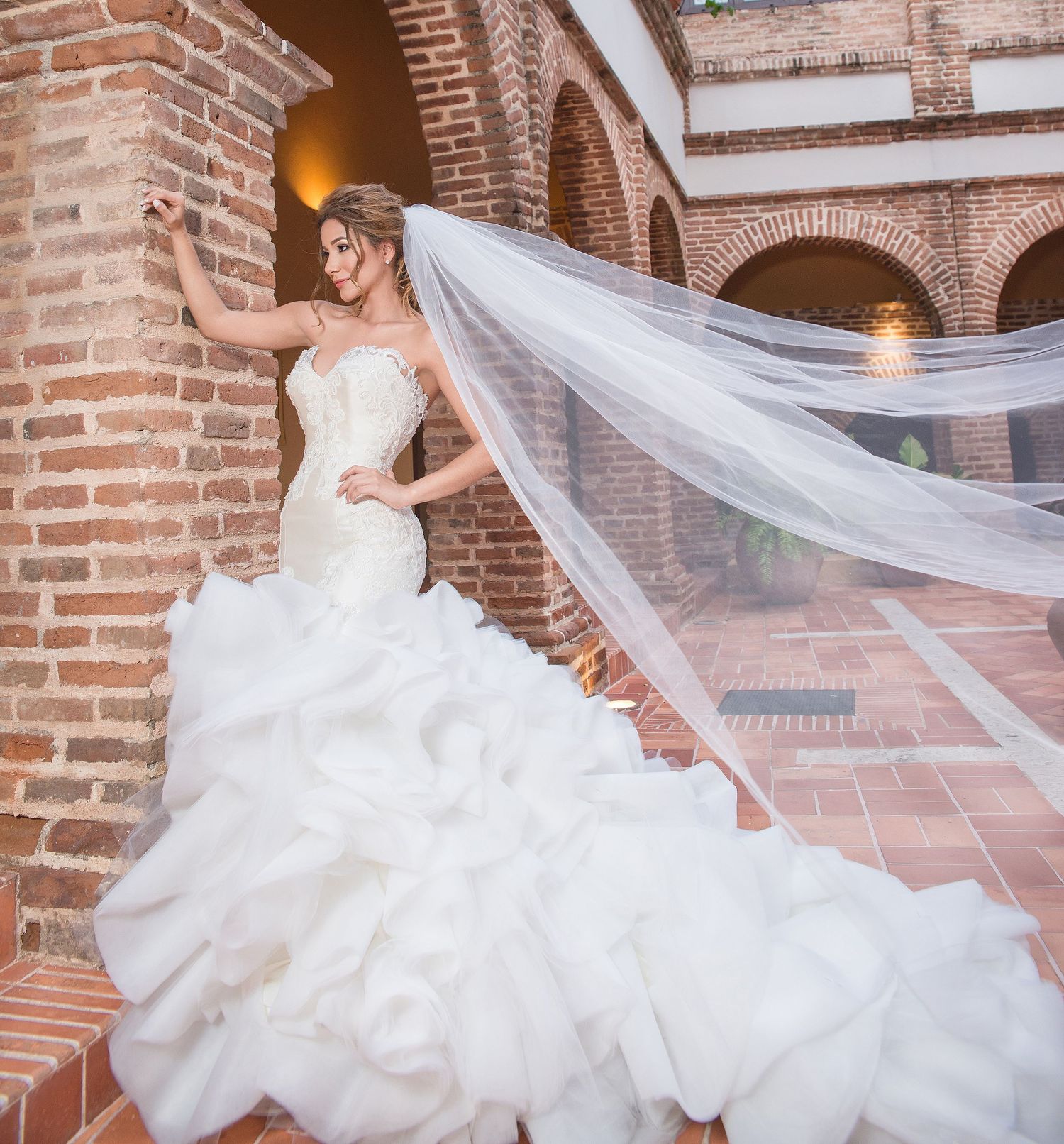 A flowing white wedding gown with dramatic train and veil against a rustic brick archway in Spanish colonial style.