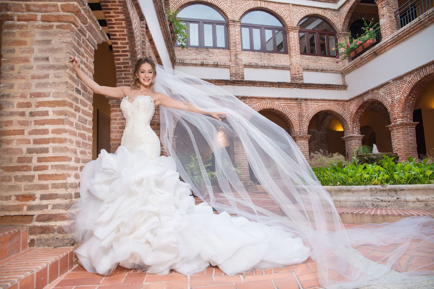 Stunning wedding dress with flowing veil billows dramatically against Spanish colonial architecture with brick arches.