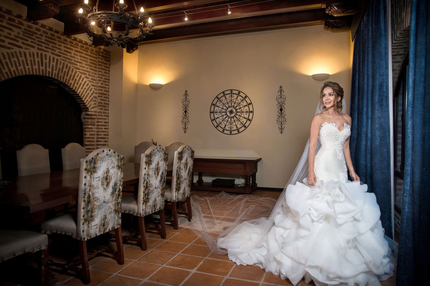 An elegant wedding dress showcased in a room with brick archways, ornate chairs and decorative wall accents.