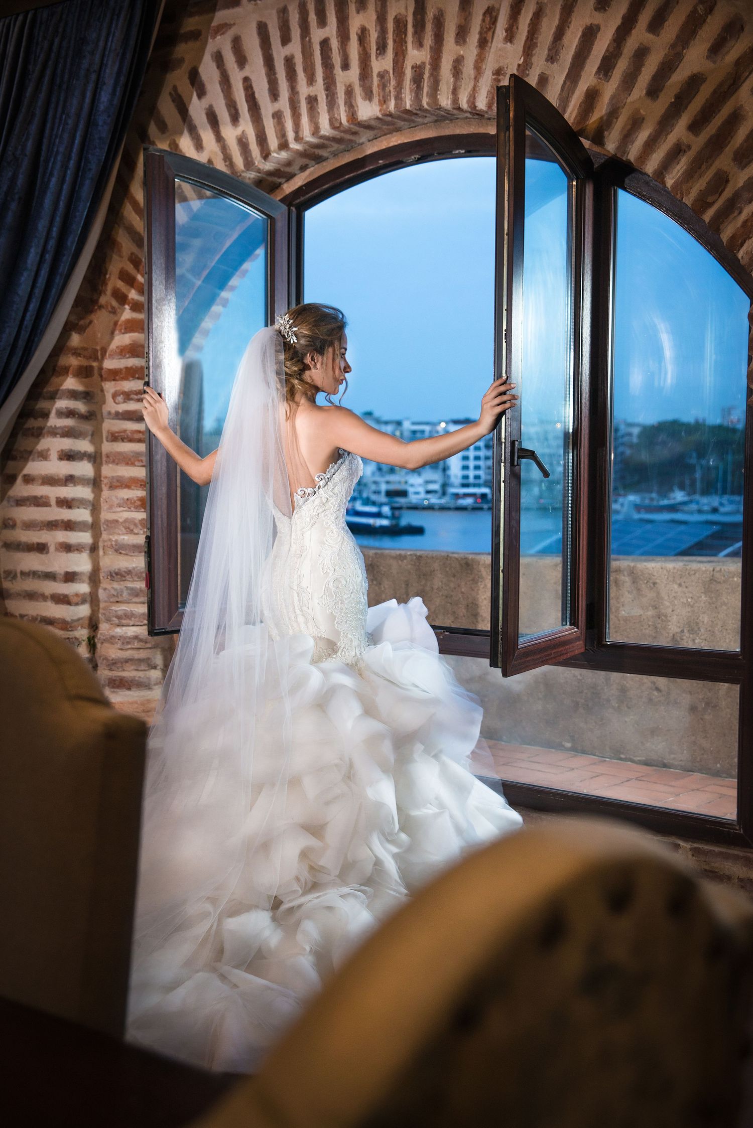 Bride in flowing white dress opens window in stone archway overlooking waterfront view.