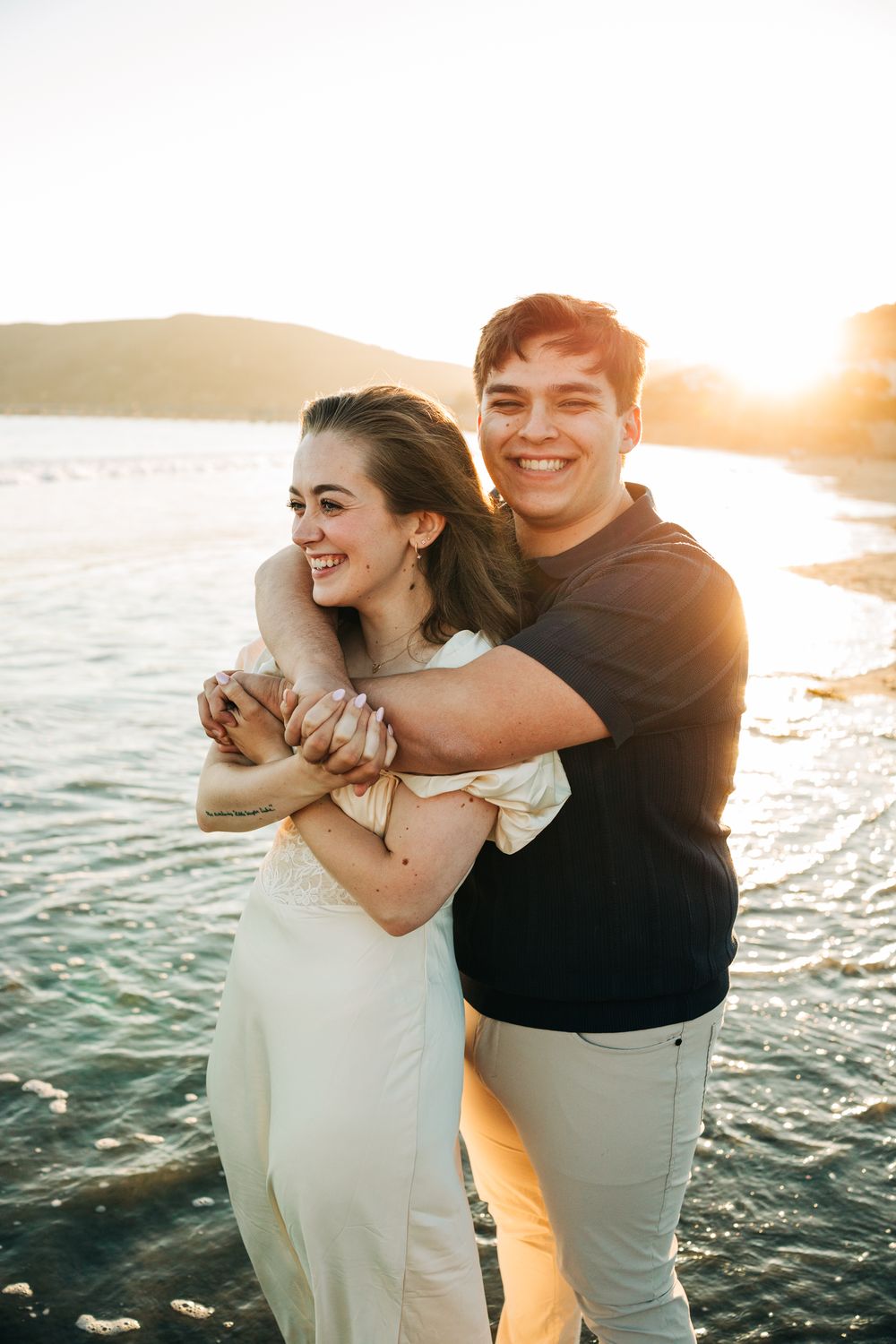 Engagement session at Pismo Beach, CA. Man hugging fiance from behind, in front of the sunset.