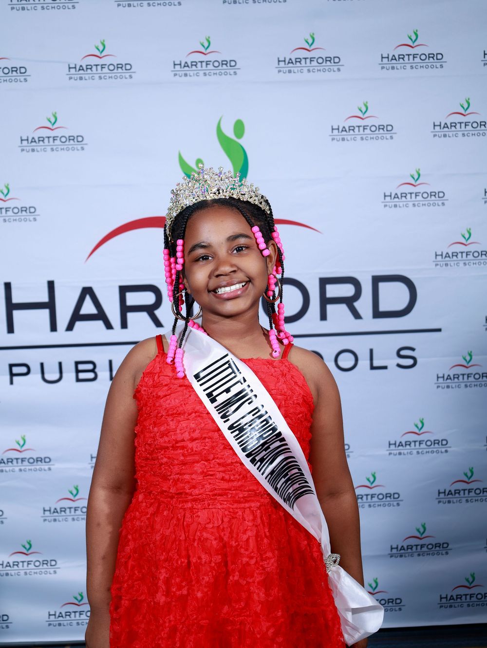 Young pageant winner in red dress wearing crown and sash at Hartford Public Schools event.