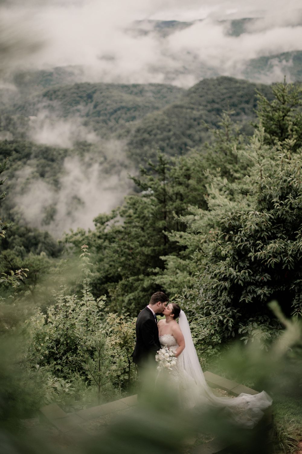 A misty mountain landscape with lush greenery creates a romantic backdrop for a couple in wedding attire.