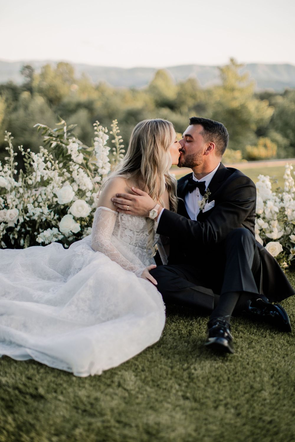 A romantic wedding couple shares intimate kisses while sitting together on grassy hillside at sunset. Nichlos Heir