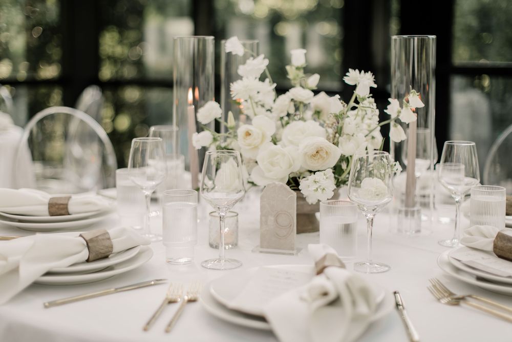 Elegant wedding reception table settings with white floral centerpieces and crystal glassware in a bright venue.