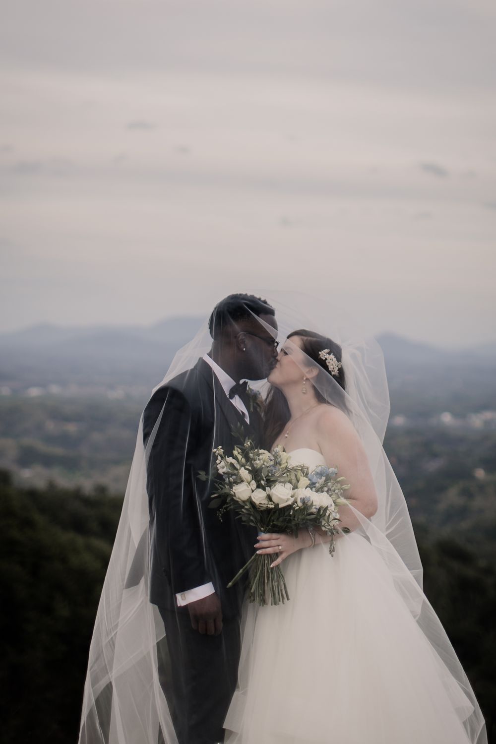 A romantic wedding couple shares a kiss on a hilltop with a misty mountain landscape in the background.