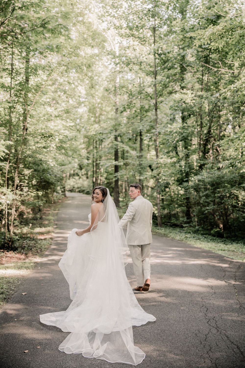A couple shares a romantic moment on a tree-lined path as the bride's flowing white dress catches the sunlight. RT LODGE