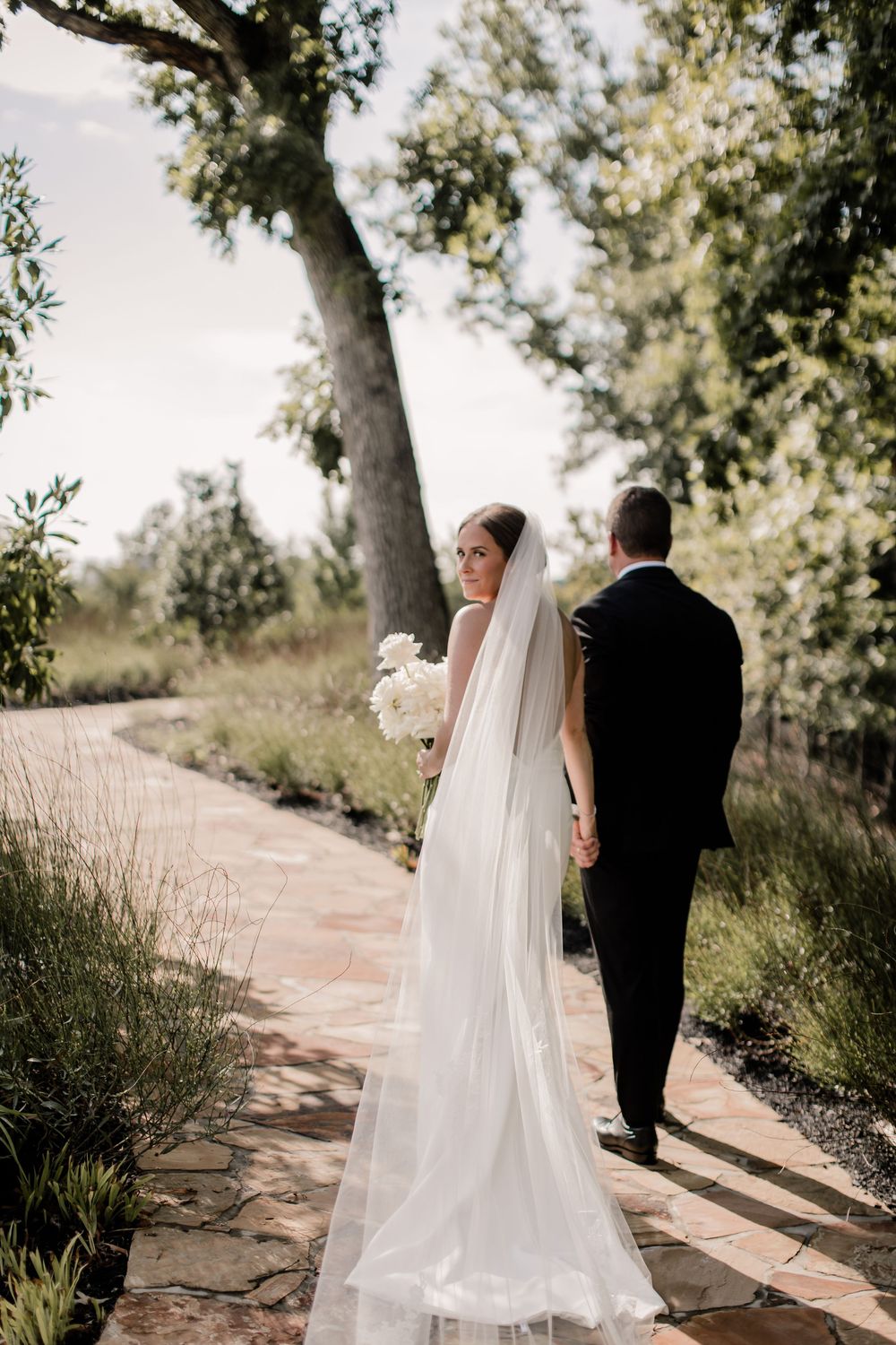 A couple in wedding attire walks along a sunlit garden path surrounded by lush greenery and trees.