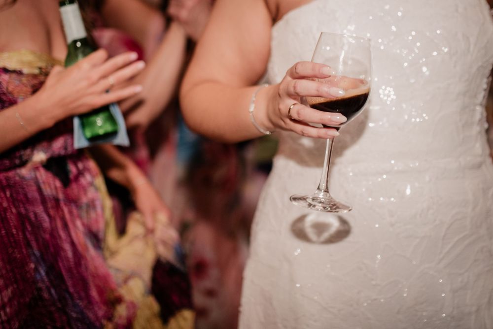 A hand in a white wedding dress holds a glass of red wine at a celebration party.