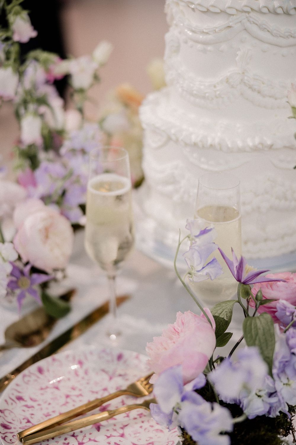 Elegant white wedding cake with delicate floral decorations, champagne glasses and pastel purple and pink flower arrangements.