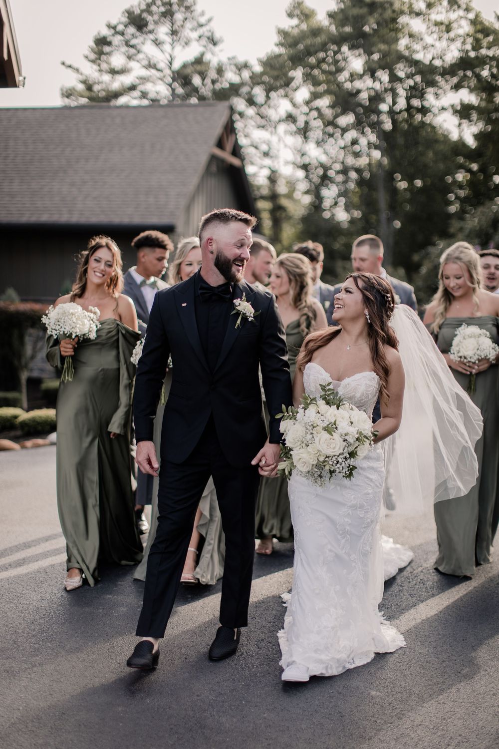 Wedding party walking down paved road with bride in white dress and groom in black suit at outdoor celebration.