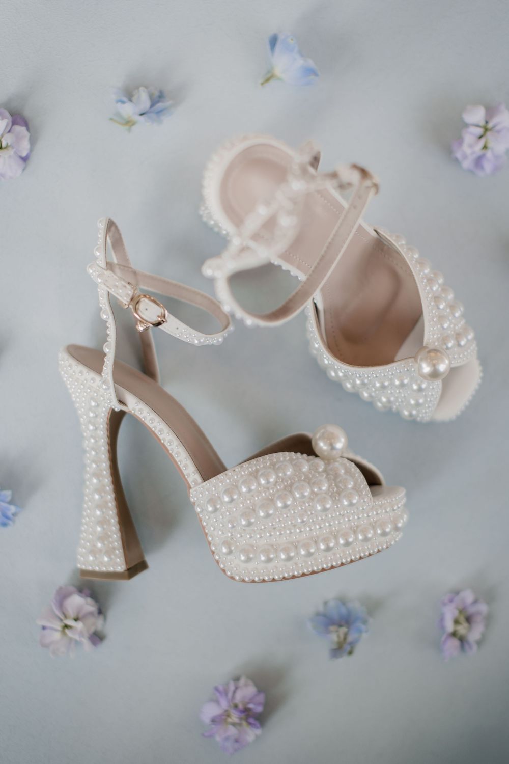 Elegant pearl-studded bridal heels with ankle straps displayed among delicate purple flowers on a light gray background.