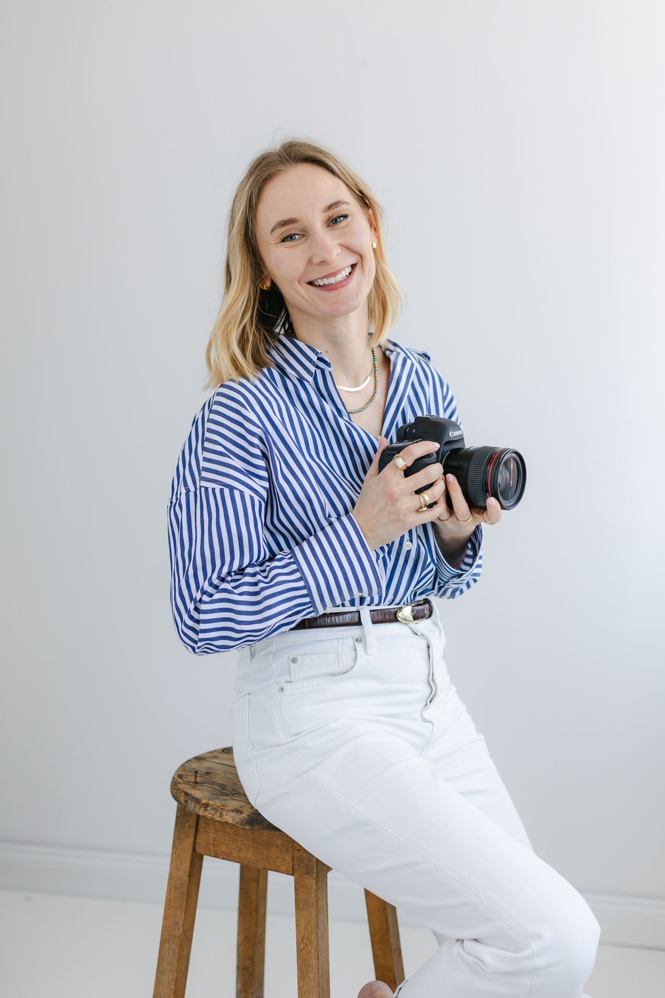 Smiling blonde female photographer sitting on a wooden stool holding a DSLR camera in a white studio.