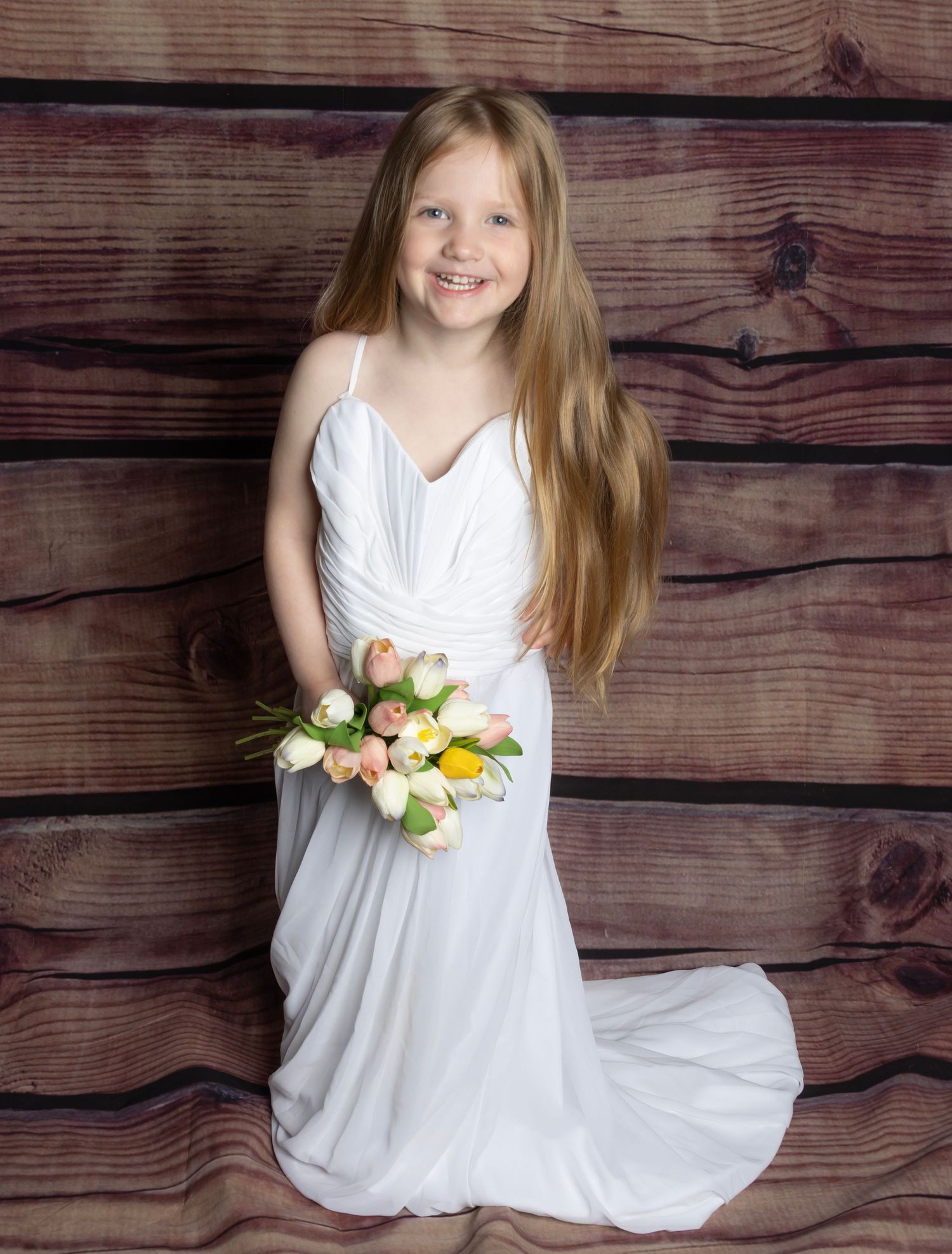 Young girl in wedding dress holding colorful tulip bouquet, smiling in front of rustic wood backdrop. Salem County Children Photographer