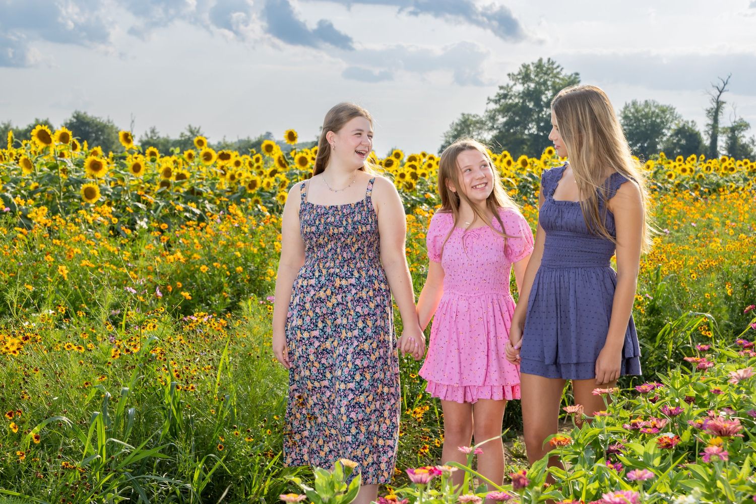 Three girls in colorful dresses smiling in a vibrant sunflower field on a sunny day. Salem and Gloucester County Children Portraits